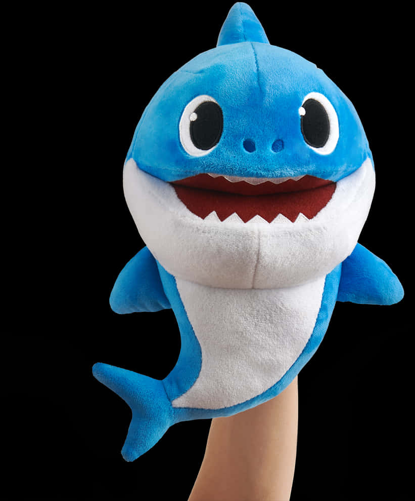 baby shark 桌布 壁紙 — 一隻藍白色毛絨嬰兒鯊魚手偶，有大眼睛和微笑的嘴巴，戴在手上，背景為黑色。