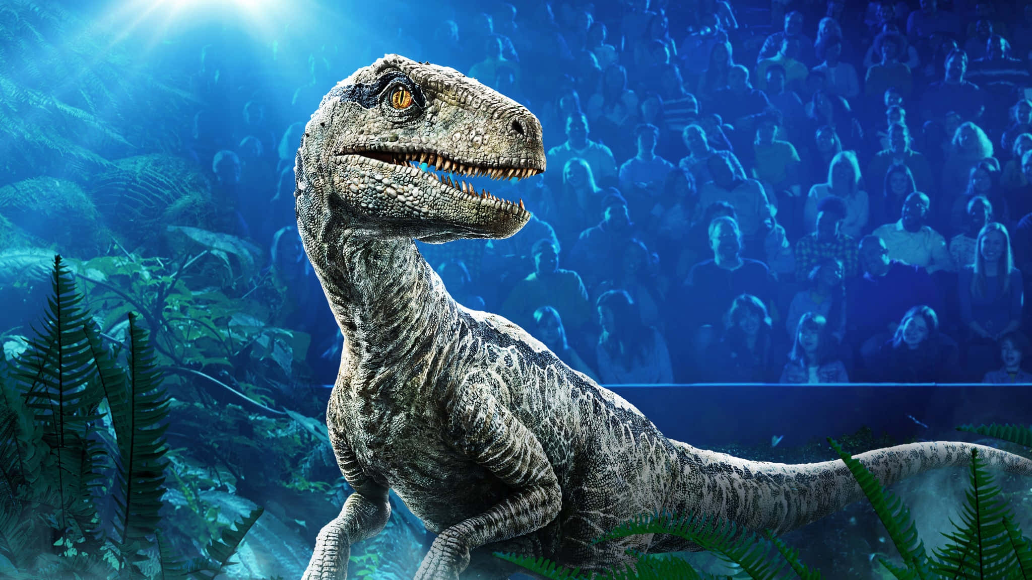 Jurassic World 桌布 壁紙 — 一隻逼真的速龍站在史前蕨類植物環境中，藍色舞台燈光下，背景有模糊的觀眾觀看。