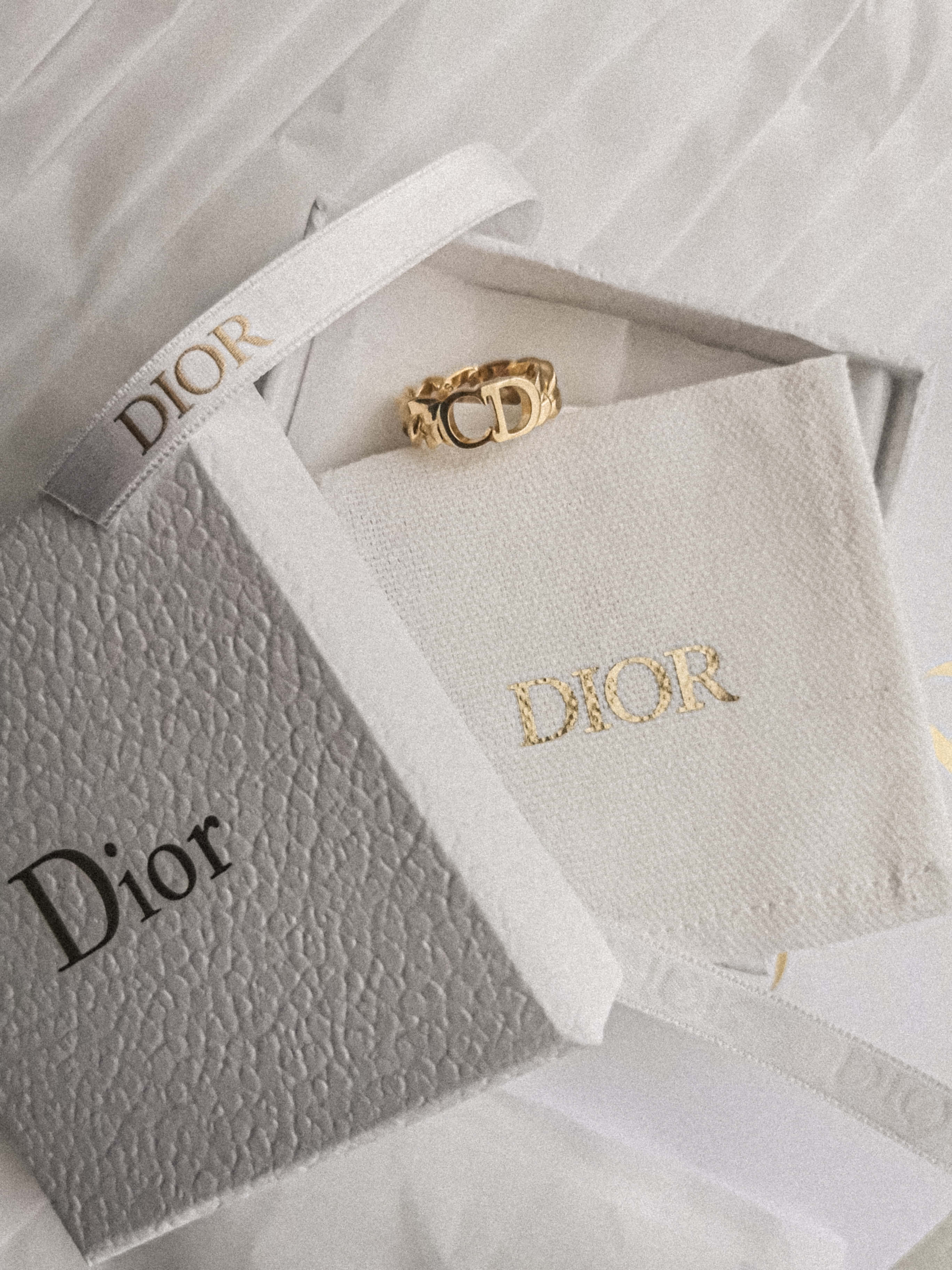 Pink Dior 桌布 壁紙 — 一隻金色Dior戒指優雅地放置在白色品牌盒子中，內襯有紋理布料和絲帶。