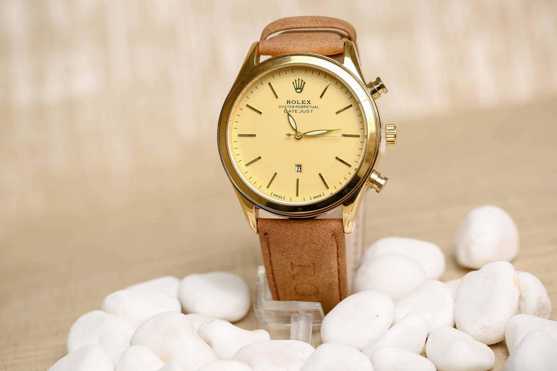 Rolex 桌布 壁紙 — 一隻金色勞力士Oyster Perpetual手錶搭配棕色皮帶，優雅地放置在光滑的白色石頭上，背景為柔和的米色。