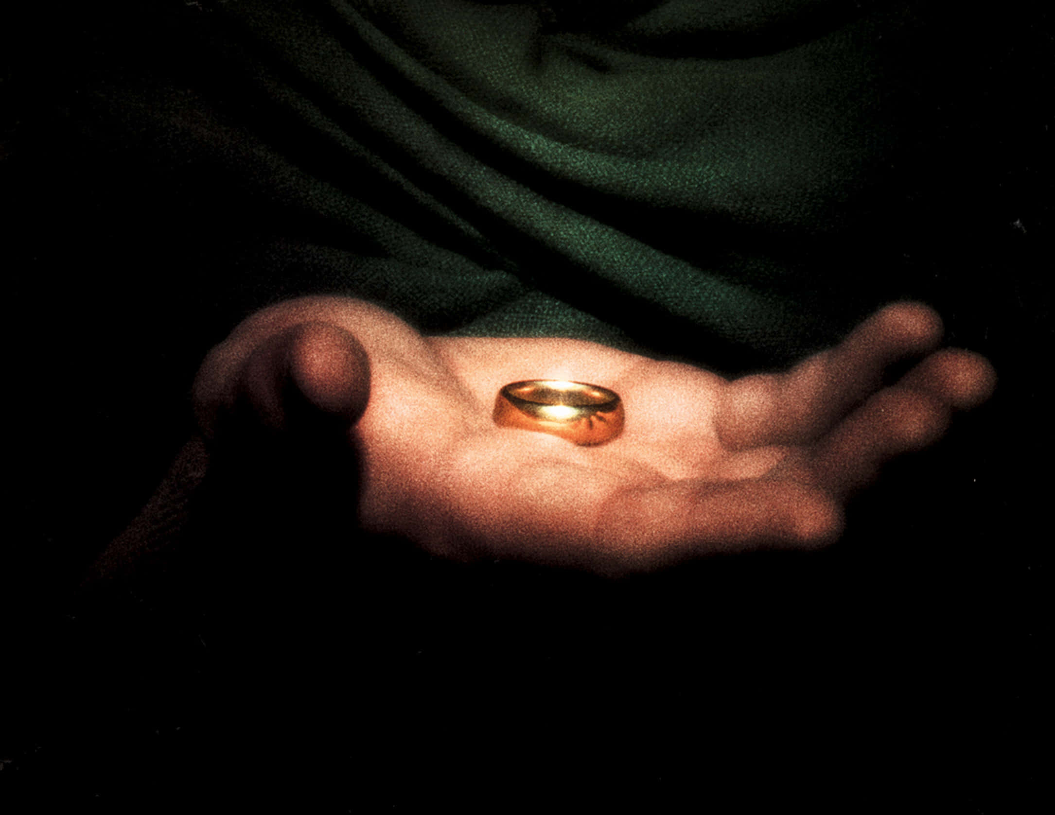 Ring 桌布 壁紙 — 一隻金色戒指在伸出的手掌中心溫暖地發光，背景是深綠色布料。