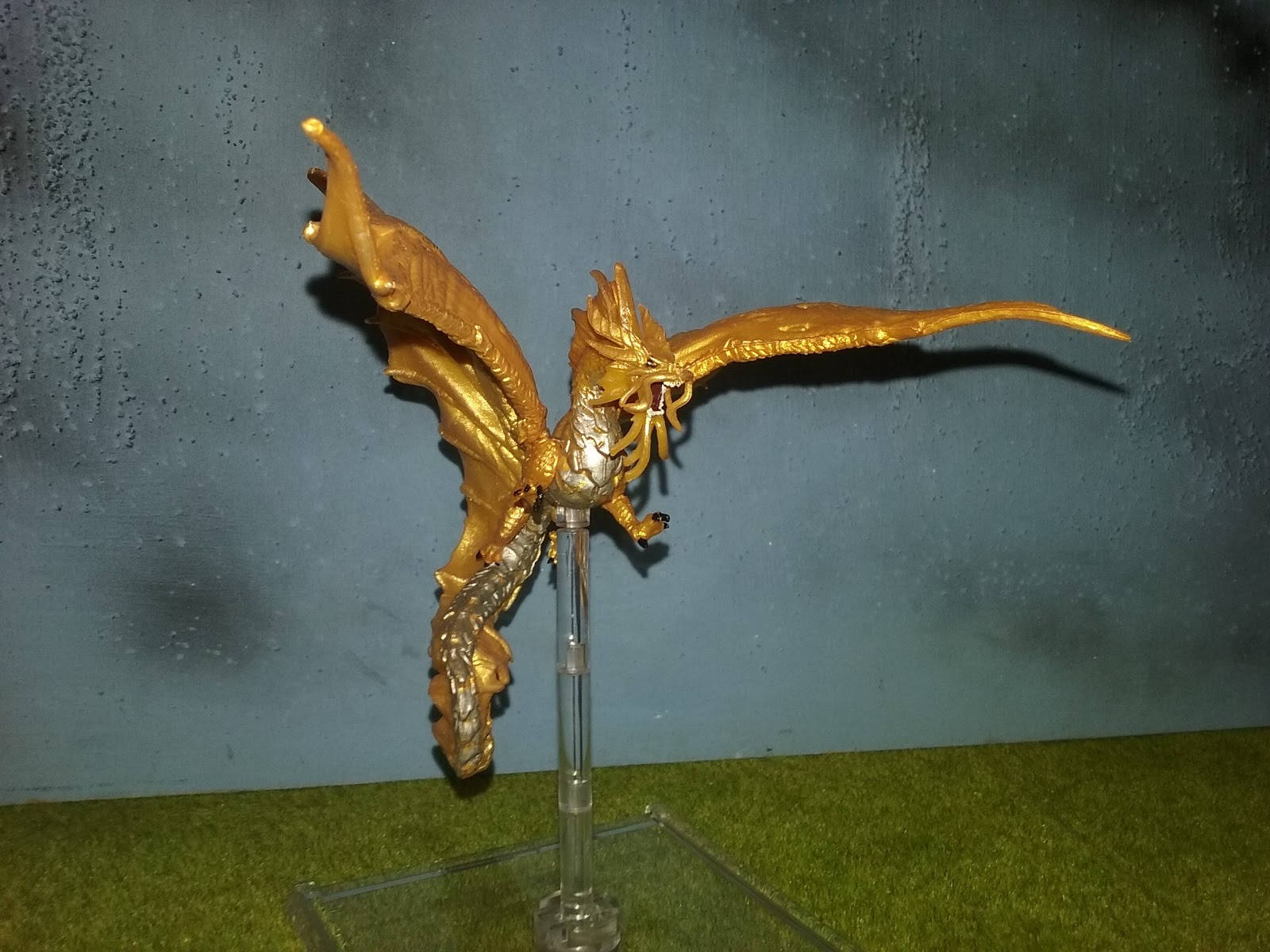 Gold Dragon 桌布 壁紙 — 一隻金色龍雕像展開翅膀，棲息在透明支架上，背景是帶紋理的藍色牆壁和綠色草地。