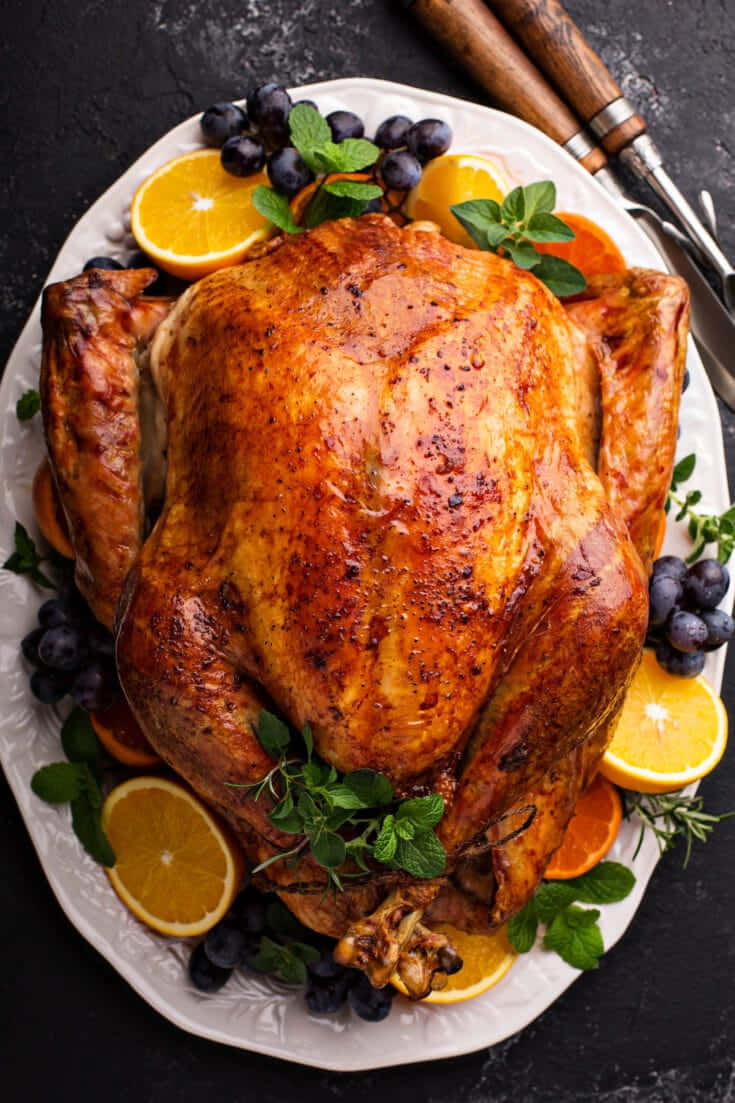 Turkey Happy Thanksgiving 桌布 壁紙 — 一隻金黃色的烤火雞，飾以柑橘片、葡萄和新鮮香草，優雅地擺放在白色托盤上，背景為深色鄉村風格。