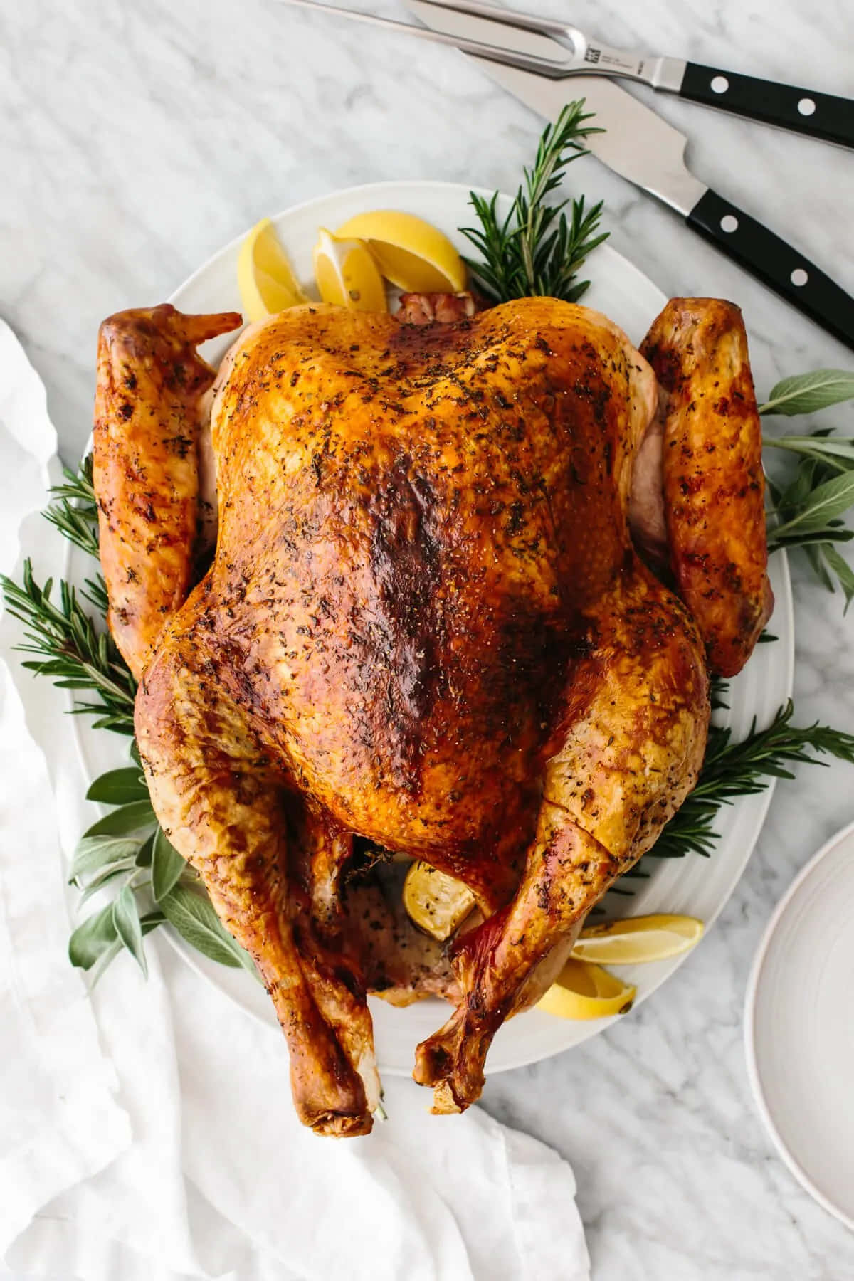 Turkey Happy Thanksgiving 桌布 壁紙 — 一隻金黃色的烤火雞，飾以迷迭香和檸檬角，放在白色盤子上，置於大理石檯面。