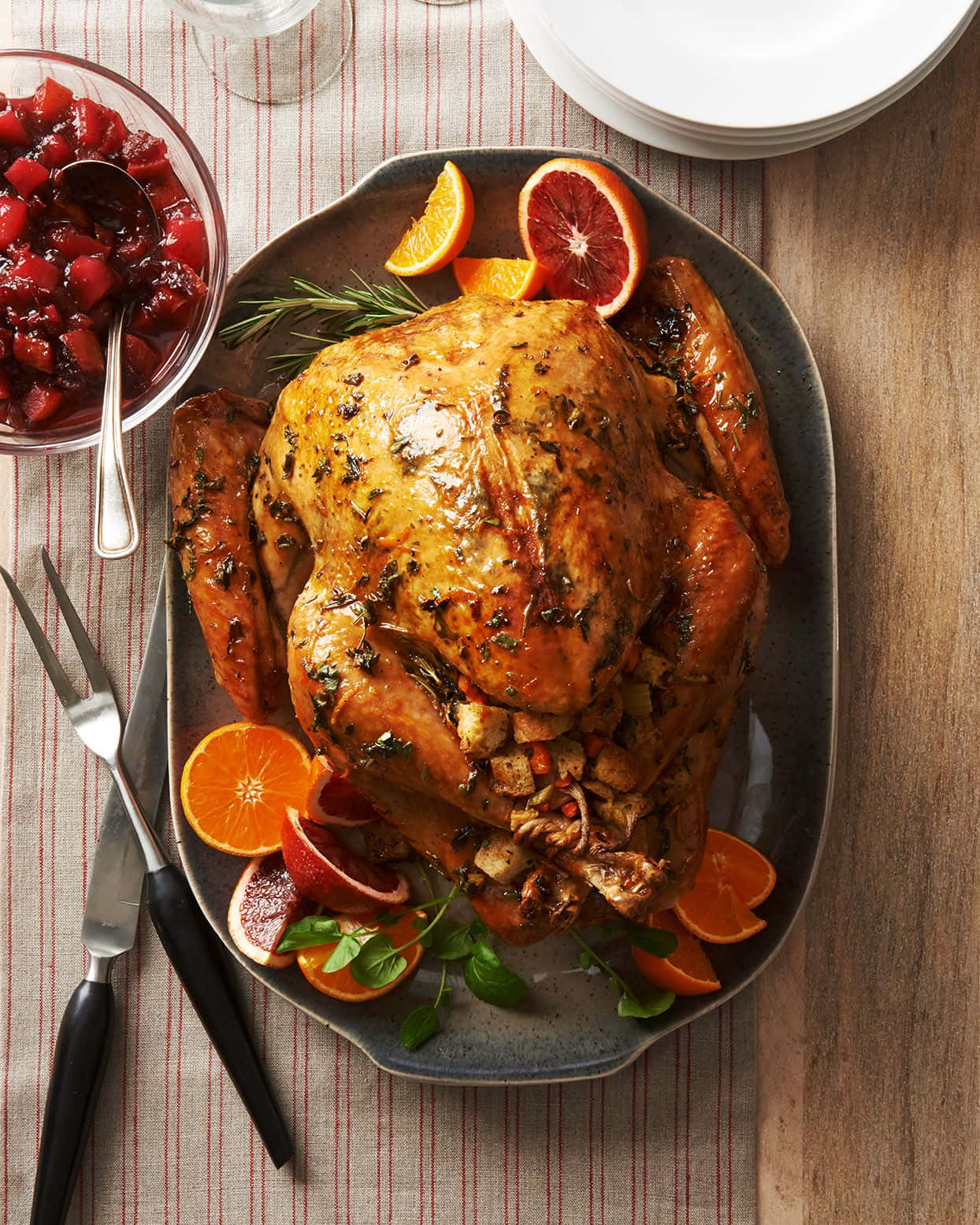 Turkey Happy Thanksgiving 桌布 壁紙 — 一隻金黃色的烤火雞，飾以香草和柑橘片，放在盤子上，旁邊有蔓越莓醬和餐具。
