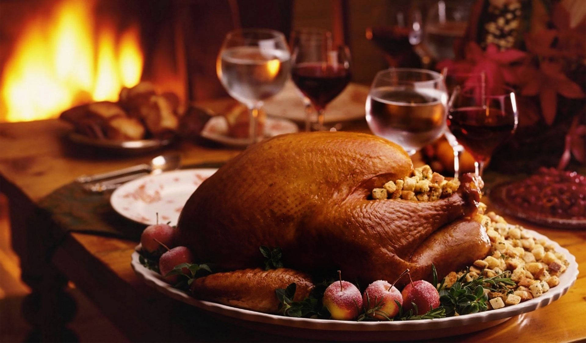 Thanksgiving 桌布 壁紙 — 一隻金黃色的烤火雞作為節日餐桌的中心裝飾，背景是酒杯和噼啪作響的壁爐。