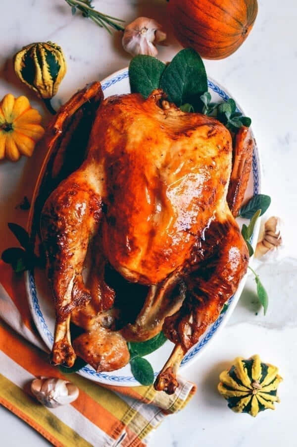 Turkey Happy Thanksgiving 桌布 壁紙 — 一隻金黃色的烤火雞在花紋盤子上閃閃發光，周圍環繞著秋季南瓜、大蒜和鼠尾草葉。
