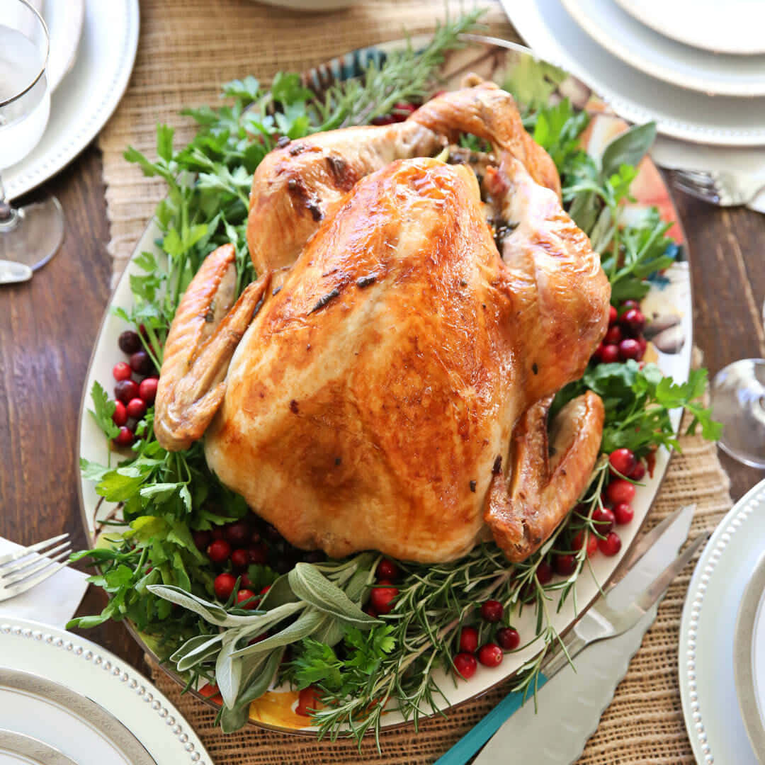 Turkey Happy Thanksgiving 桌布 壁紙 — 一隻金黃色的烤火雞放在托盤上，周圍是新鮮香草和蔓越莓，置於鄉村風木桌上。