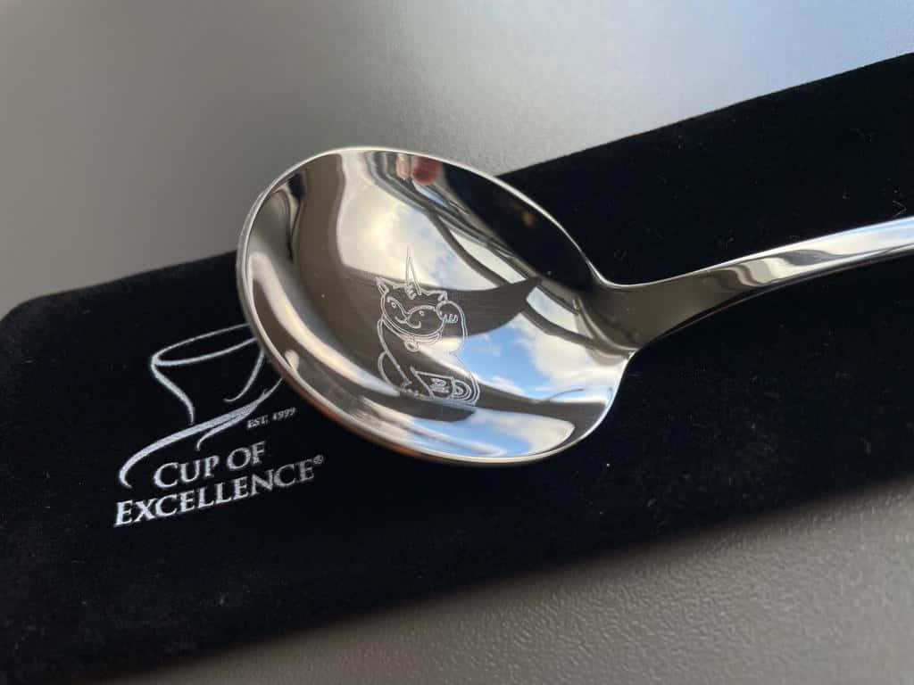 kitchen 桌布 壁紙 — 一隻閃亮的銀湯匙，上面刻有貓圖案，放在印有「Cup of Excellence」品牌的黑色天鵝絨袖套上。