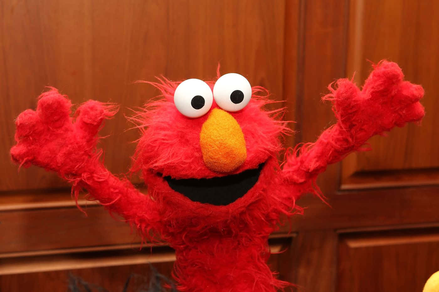Elmo 桌布 壁紙 — 一隻開心的紅色毛絨公仔，有大白色眼睛和橙色鼻子，在木背景前開心地揮手。