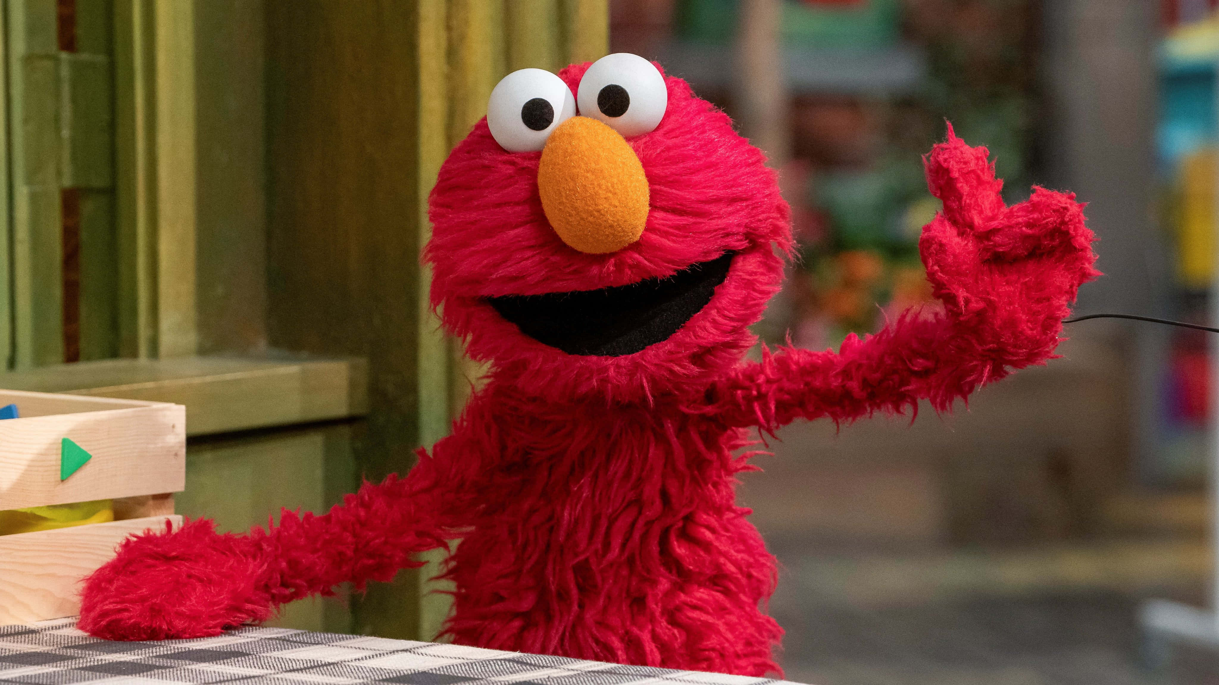 Elmo 桌布 壁紙 — 一隻開心的紅色毛絨玩偶，有大眼睛和橙色鼻子，在格子桌後面開心地揮手。
