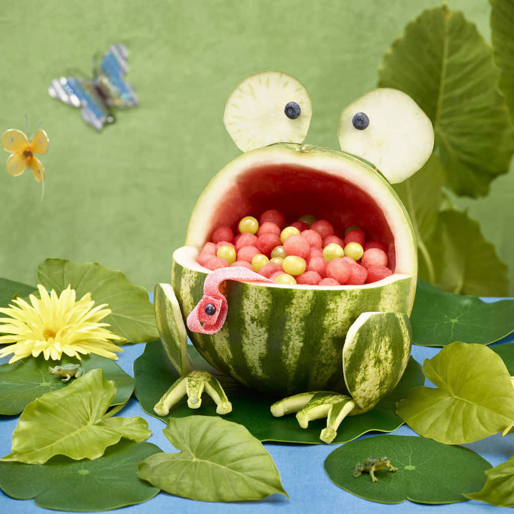 Watermelon 桌布 壁紙 — 一隻雕刻的西瓜青蛙，有藍莓眼睛和裝滿糖果的嘴巴，坐在荷葉和蝴蝶之間。