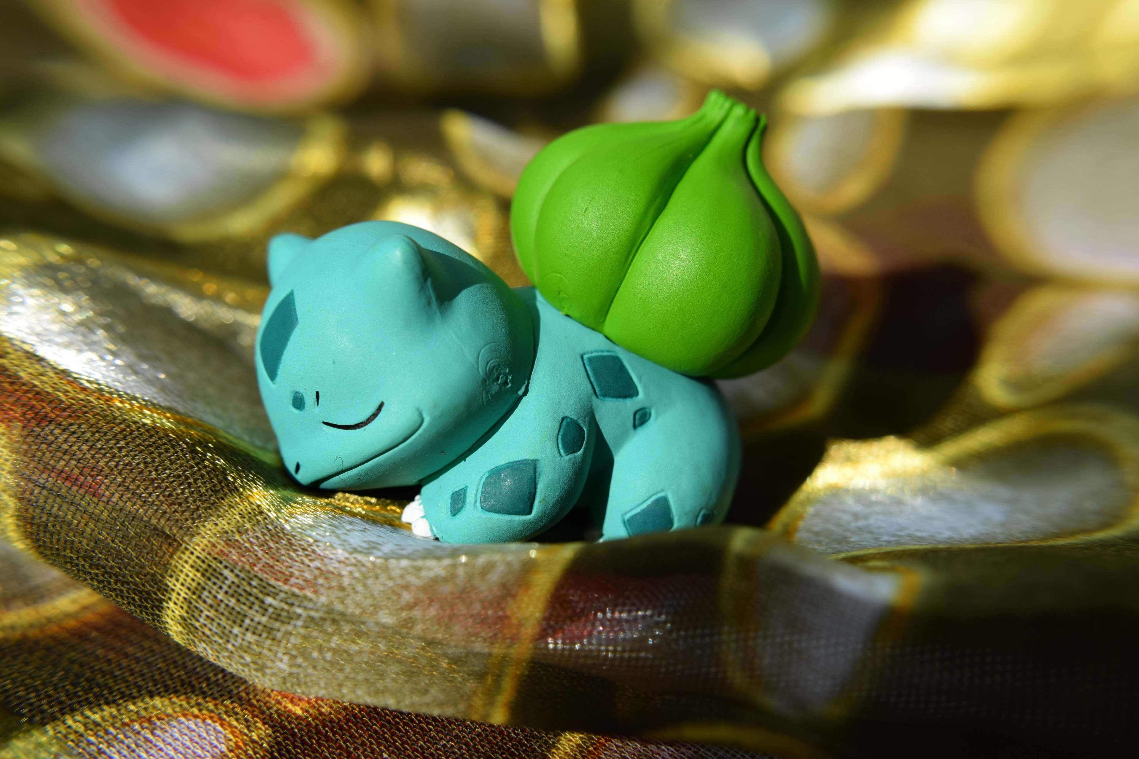 Pokemon Toys 桌布 壁紙 — 一隻青綠色的妙蛙種子公仔在帶有圖案的金色布料上平靜地休息，伴隨著柔和溫暖的燈光。