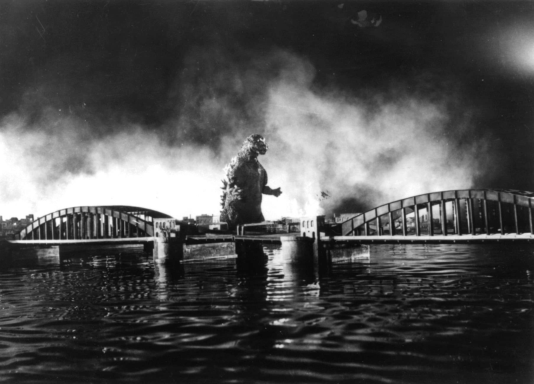Godzilla 1954 桌布 壁紙 — 一隻高大的哥斯拉站在水中，位於兩座斷橋之間，身後濃煙滾滾，形成戲劇性的黑白場景。