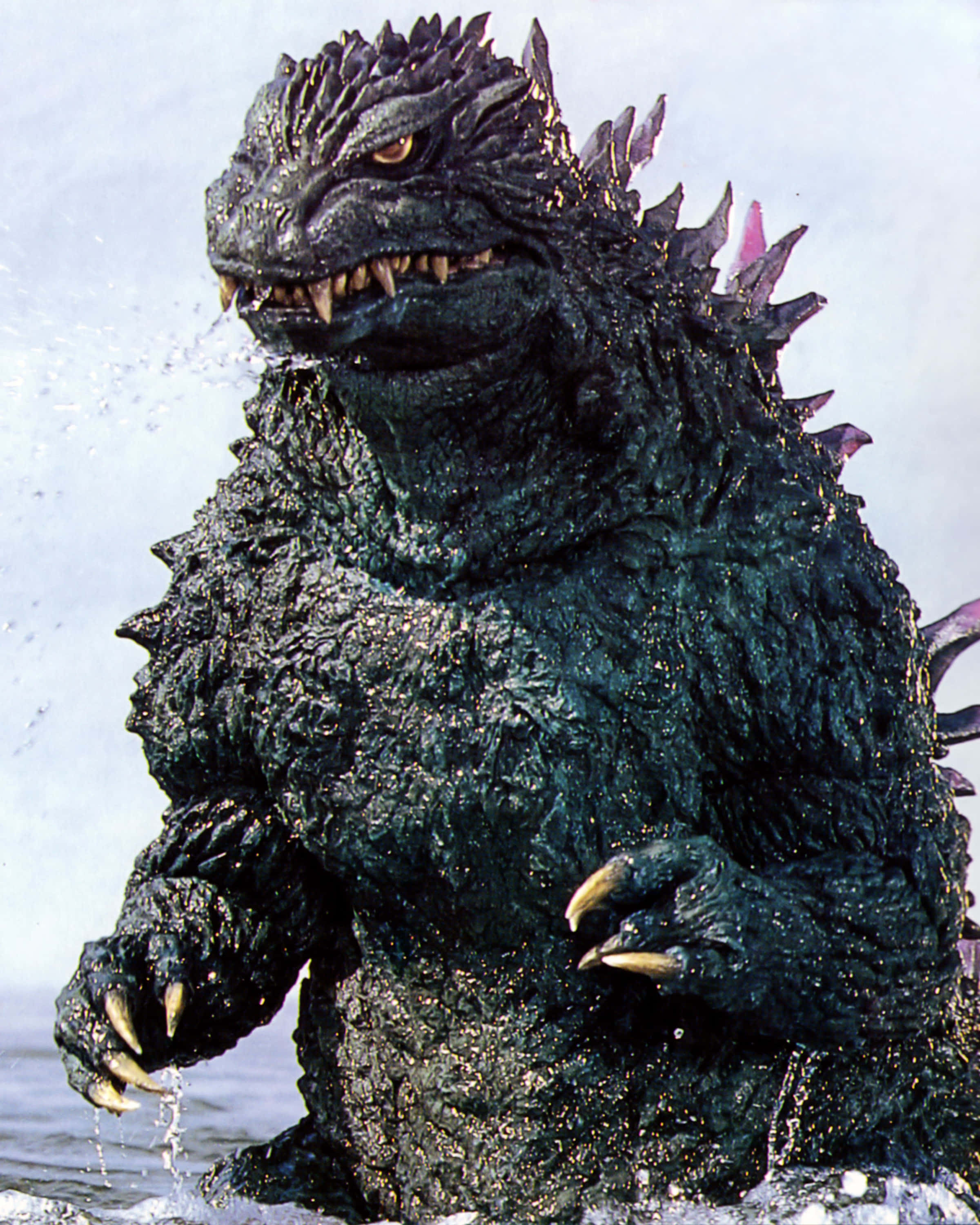 Godzilla 2000 桌布 壁紙 — 一隻高聳的哥吉拉從洶湧的海水中出現，帶有尖刺的背鰭和發光的眼睛。