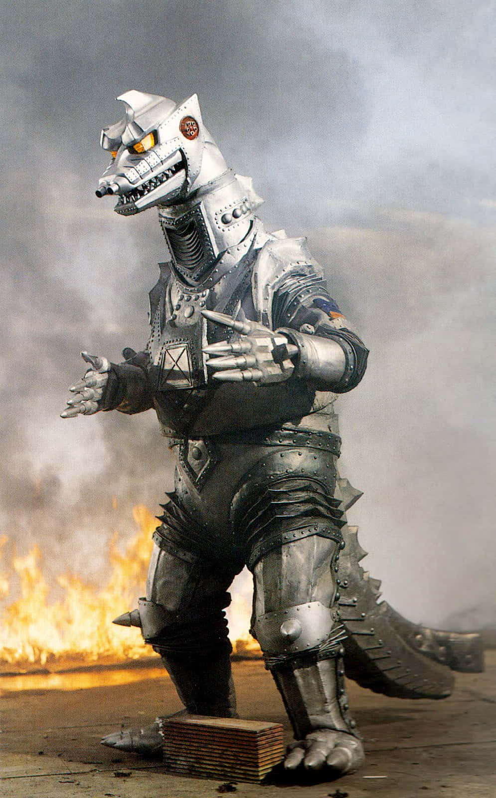 mechagodzilla 桌布 壁紙 — 一隻高聳的銀色裝甲怪獸站在烈火毀滅之中，散發著橙色發光的眼睛，身後煙霧翻騰。