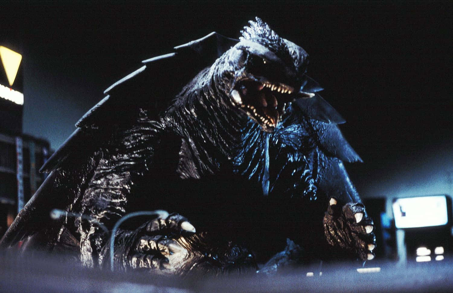 Gamera 桌布 壁紙 — 一隻高聳的黑色哥吉拉長有翅膀，在黑暗的城市夜景中，伴隨發光的城市燈光，發出威嚇的咆哮。