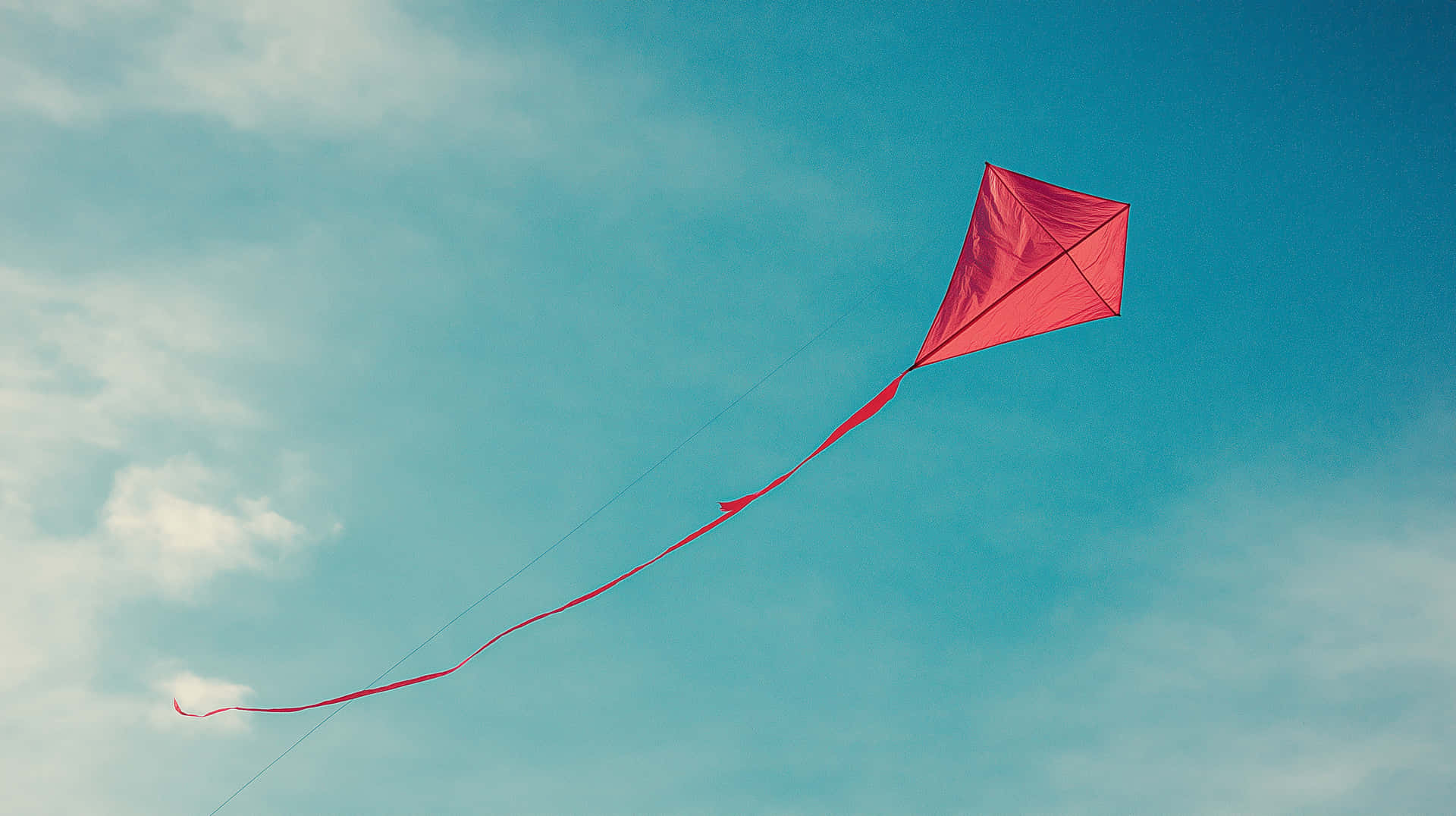 Red Diamond Kite Flying High Against a Bright Blue Sky 壁紙 — 一隻鮮紅色的菱形風箏拖著長長的尾巴，在點綴著縷縷輕雲的柔和藍天中優雅地翱翔。