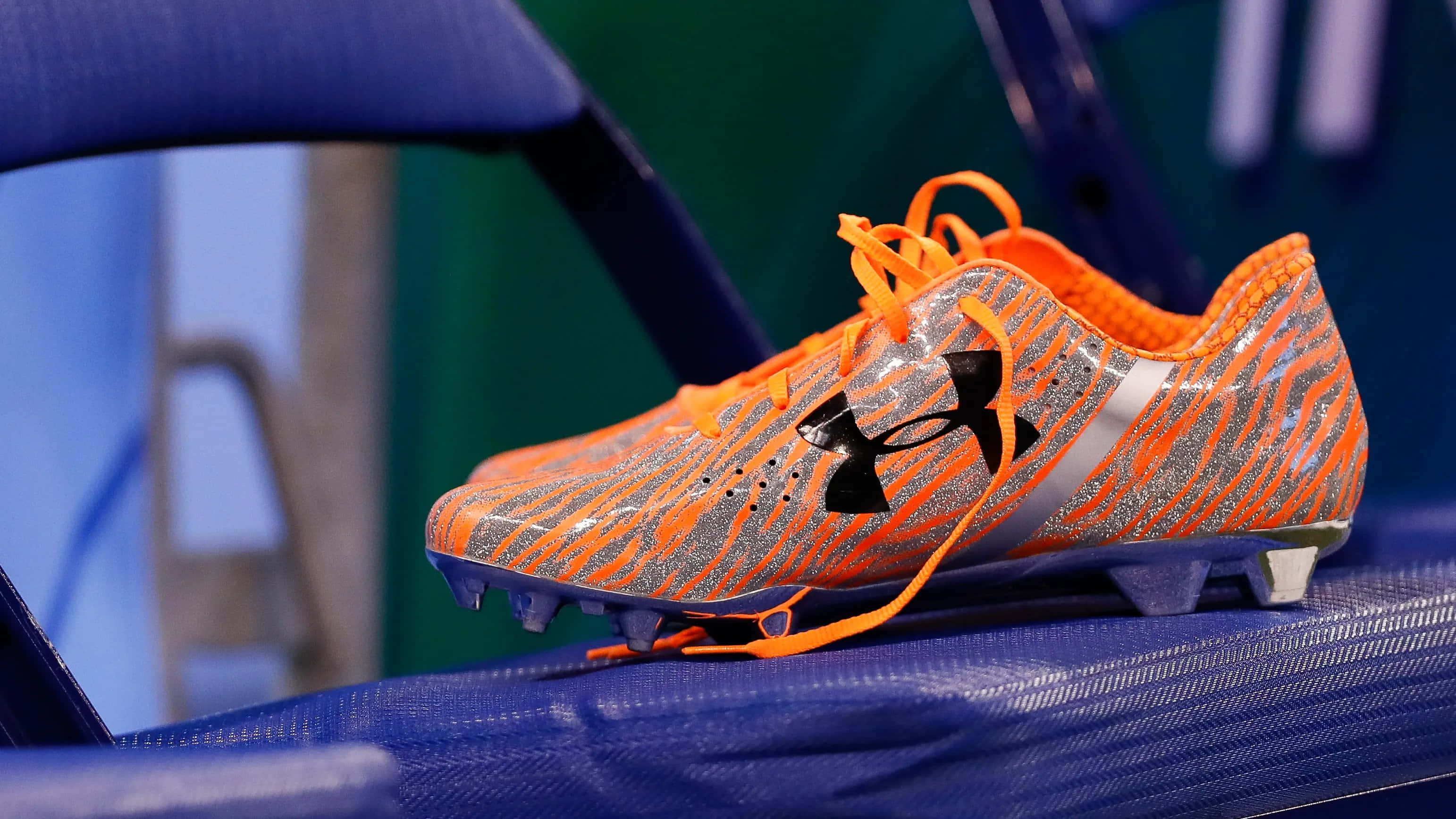 Under Armour 桌布 壁紙 — 一隻鮮豔的橙色和銀色足球鞋，帶有黑色標誌，放在藍色體育場座位上。