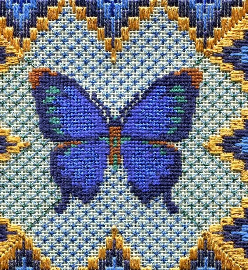 Butterfly Stitch 桌布 壁紙 — 一隻鮮豔的藍色蝴蝶，帶有綠色和橙色點綴，以十字繡縫在圖案布料背景上。