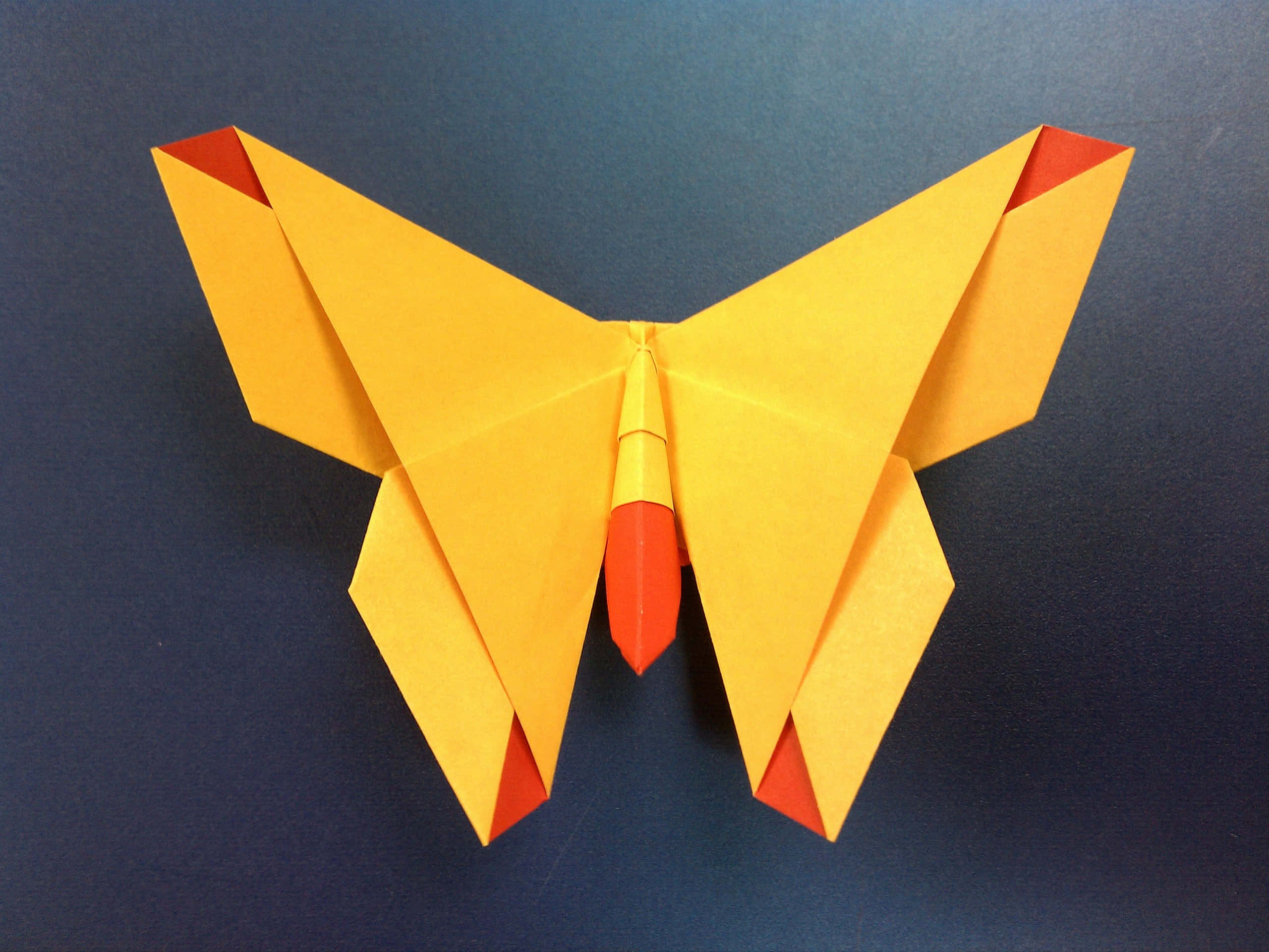 Butterfly Origami 桌布 壁紙 — 一隻鮮豔的黃色和紅色摺紙蝴蝶，帶有銳利的幾何摺痕，停留在深藍色背景上。