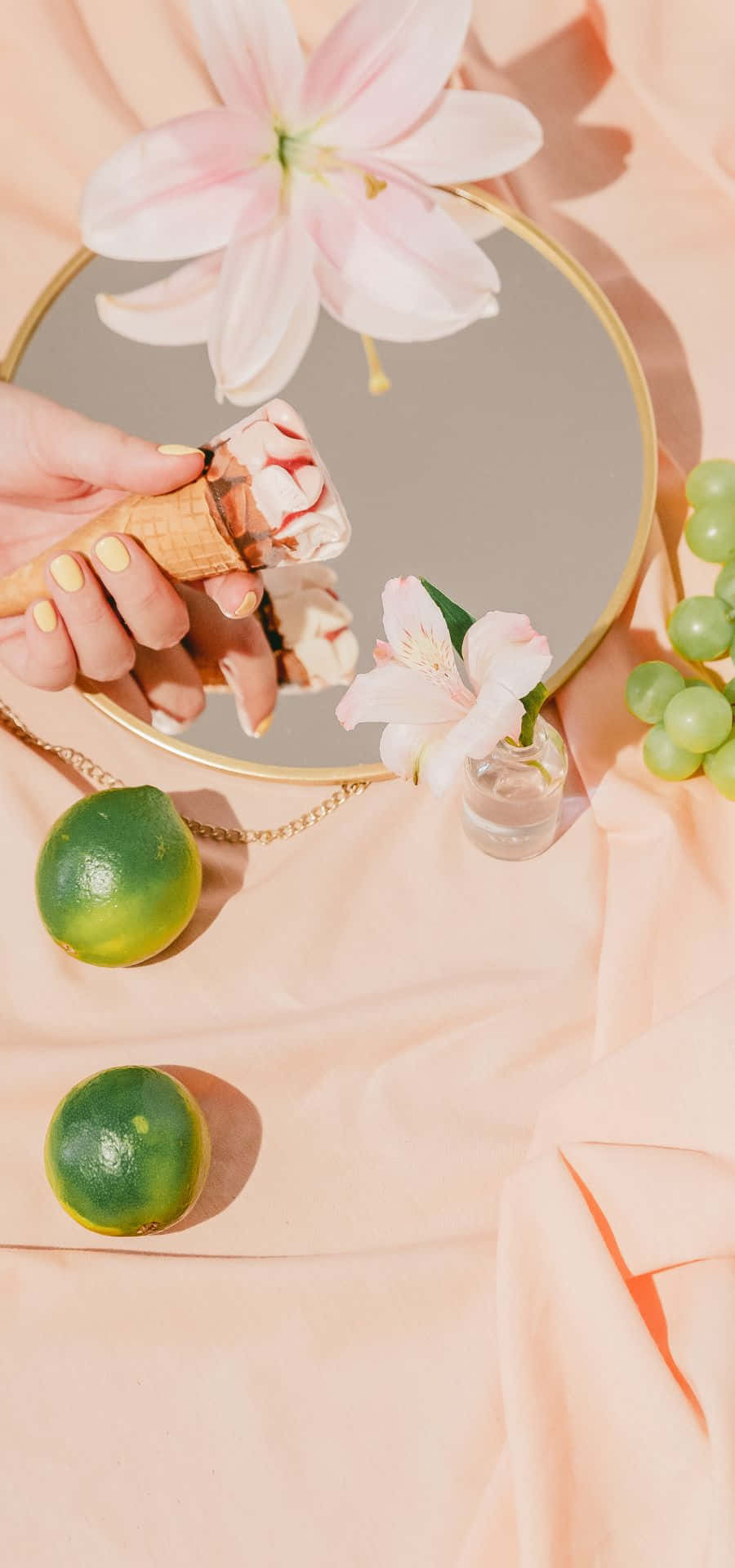 Peach Color 桌布 壁紙 — 一隻黃色指甲的手在圓鏡上方拿著冰淇淋甜筒，鏡中反射出粉紅花朵和青葡萄，下方是柔軟的桃色布料。