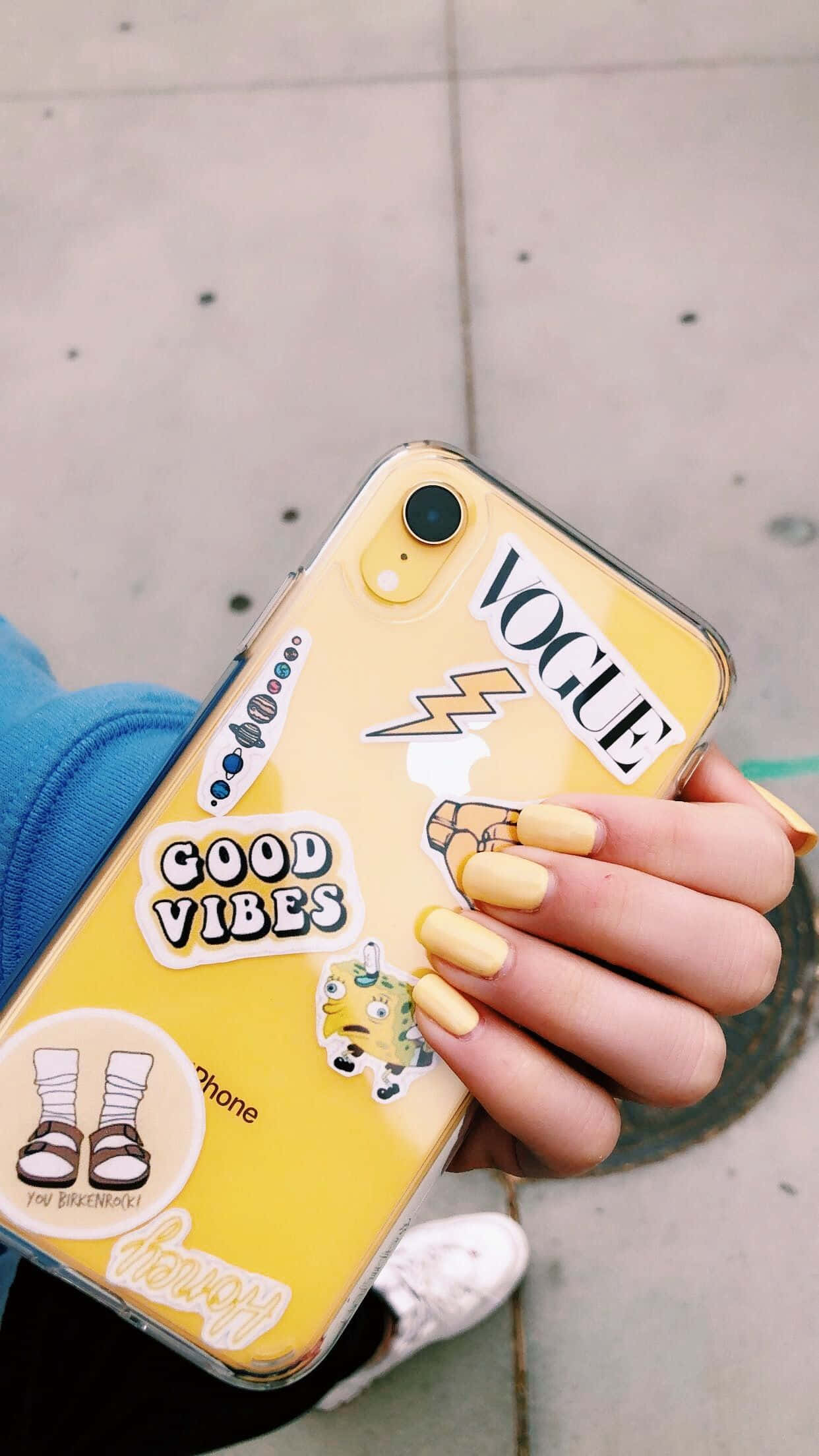 Phone Case 桌布 壁紙 — 一隻黃色指甲的手拿著一個黃色手機殼，上面貼滿了包括Vogue和Good Vibes在內的彩色貼紙，背景是行人道。