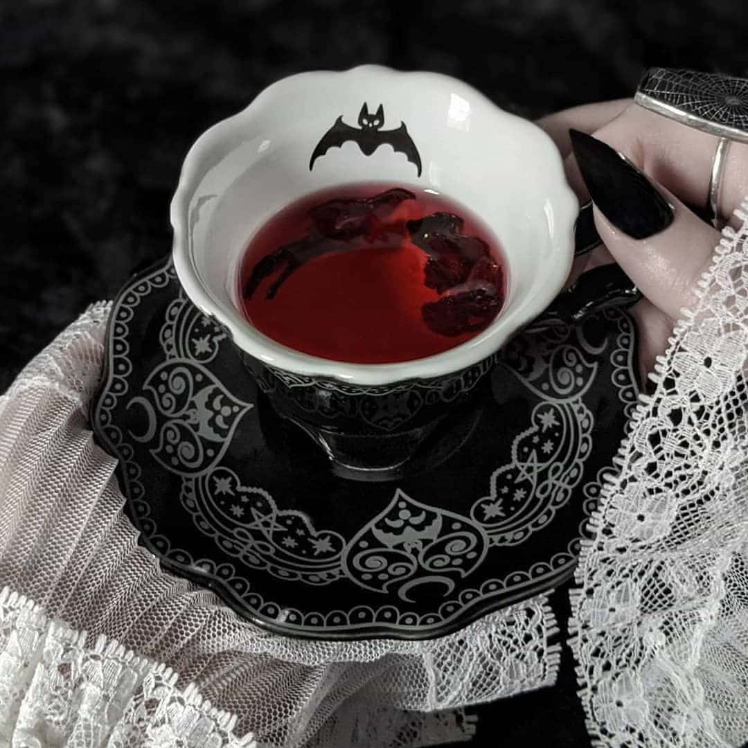 Vampire Aesthetic 桌布 壁紙 — 一隻黑色指甲的手拿著一個帶有蝙蝠圖案的白色杯子，裡面裝著紅茶，放在一個帶有蕾絲邊裝飾的黑色茶托上。