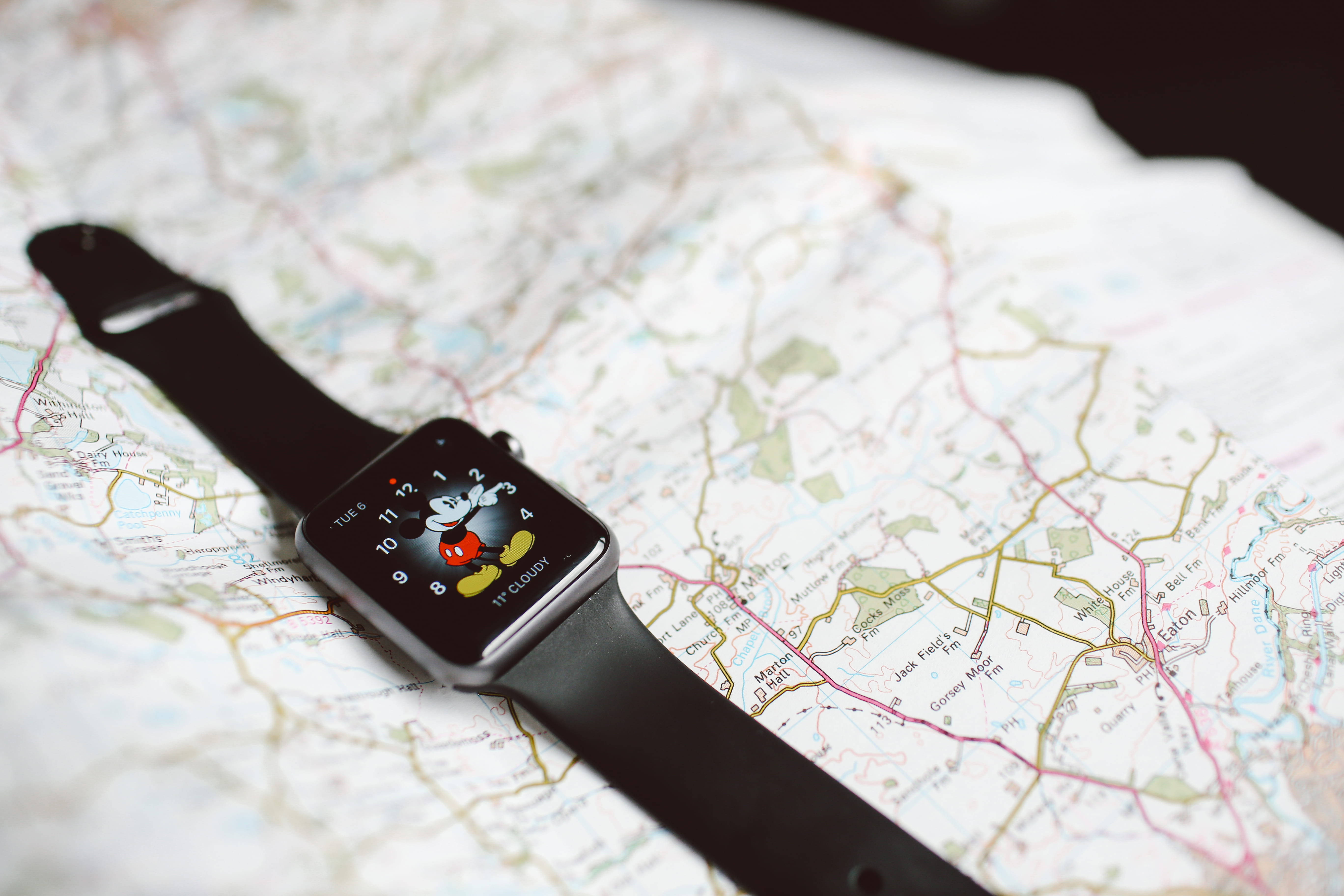 Apple Watch 動態 桌布 壁紙 — 一隻黑色智慧手錶帶有米奇老鼠錶面，放在一張顯示道路和城鎮的詳細紙質地圖上。