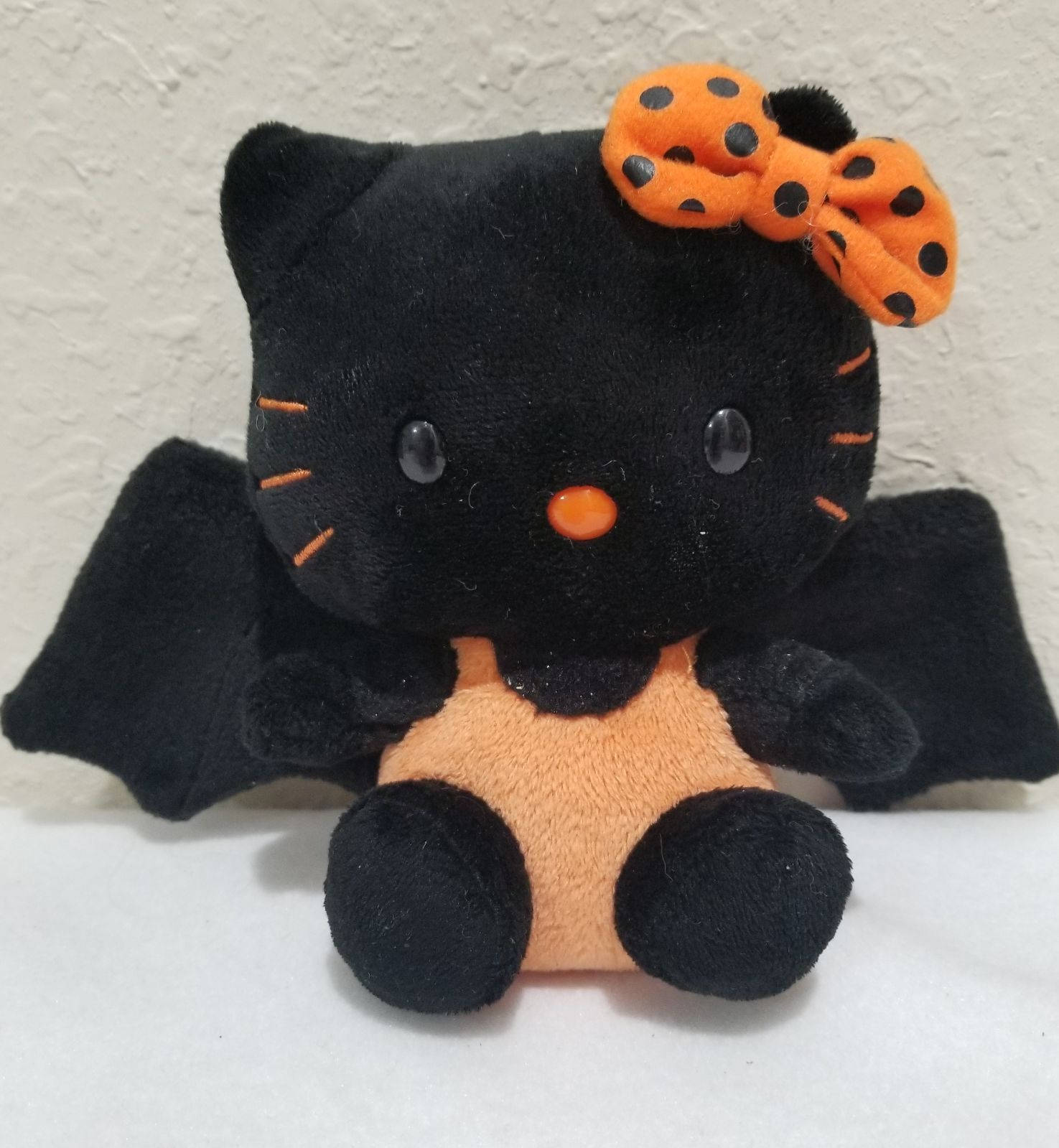 Hello Kitty Halloween 桌布 壁紙 — 一隻黑色絨毛Hello Kitty扮成蝙蝠，戴著橙色圓點蝴蝶結和橙色肚子，坐在有紋理的牆前。