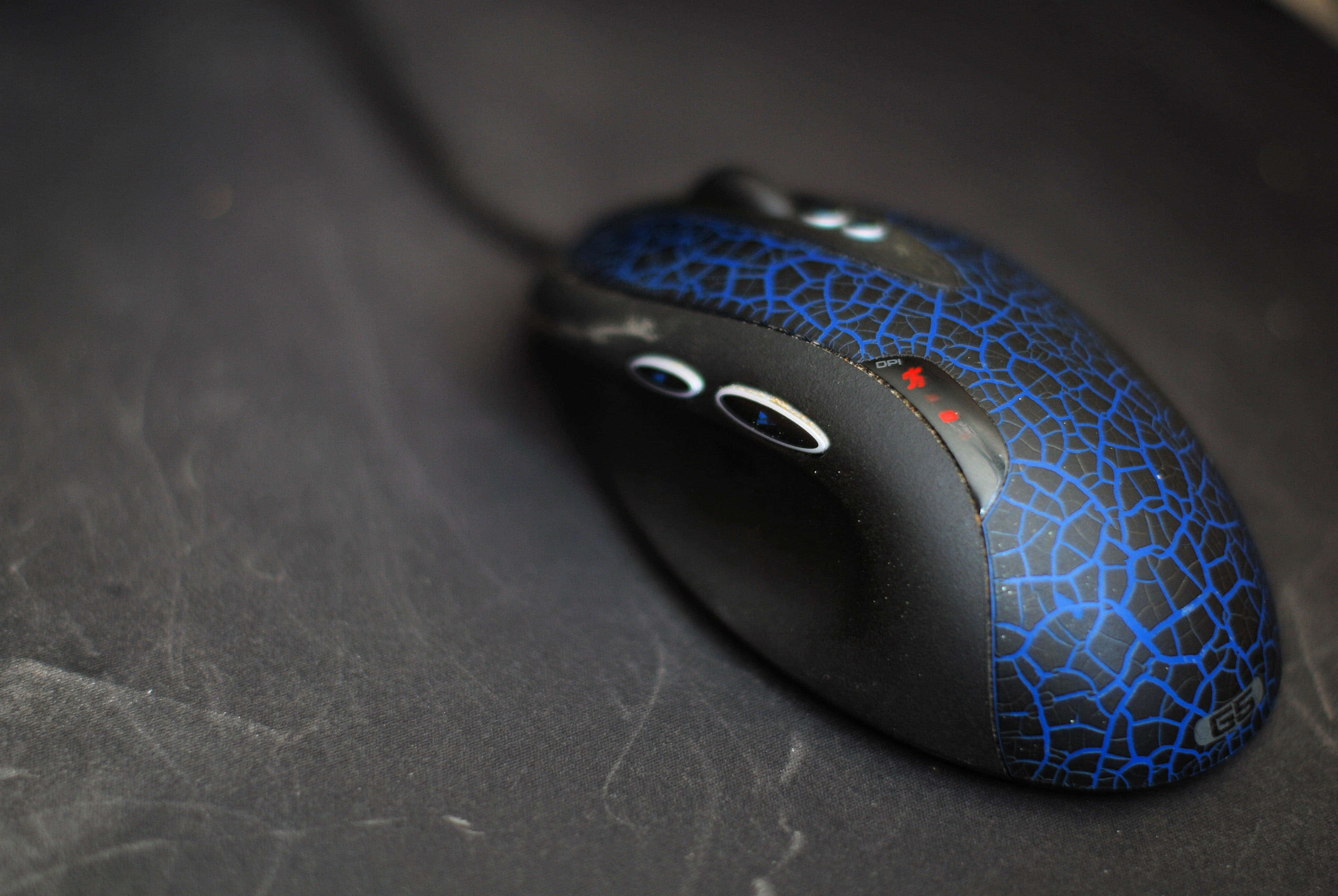 Gaming Mice 桌布 壁紙 — 一隻黑色電競滑鼠帶有藍色裂紋圖案點綴，放置在刮花的深色表面上。