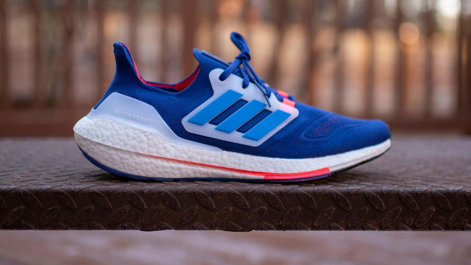 愛迪達 桌布 壁紙 — 一雙充滿活力的藍色 Adidas Ultraboost 運動鞋，帶有白色和紅色點綴，放置在紋理金屬表面上，背景溫暖模糊。