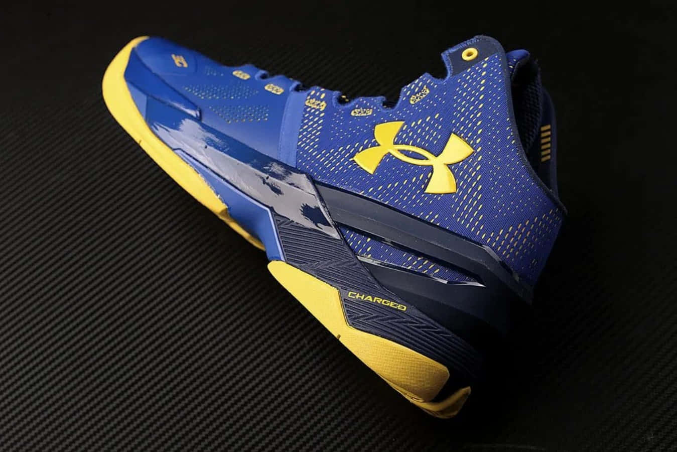 Under Armour 桌布 壁紙 — 一雙充滿活力的藍黃色 Under Armour 籃球鞋，帶有「CHARGED」品牌標誌，放置在紋理黑色碳纖維表面上。