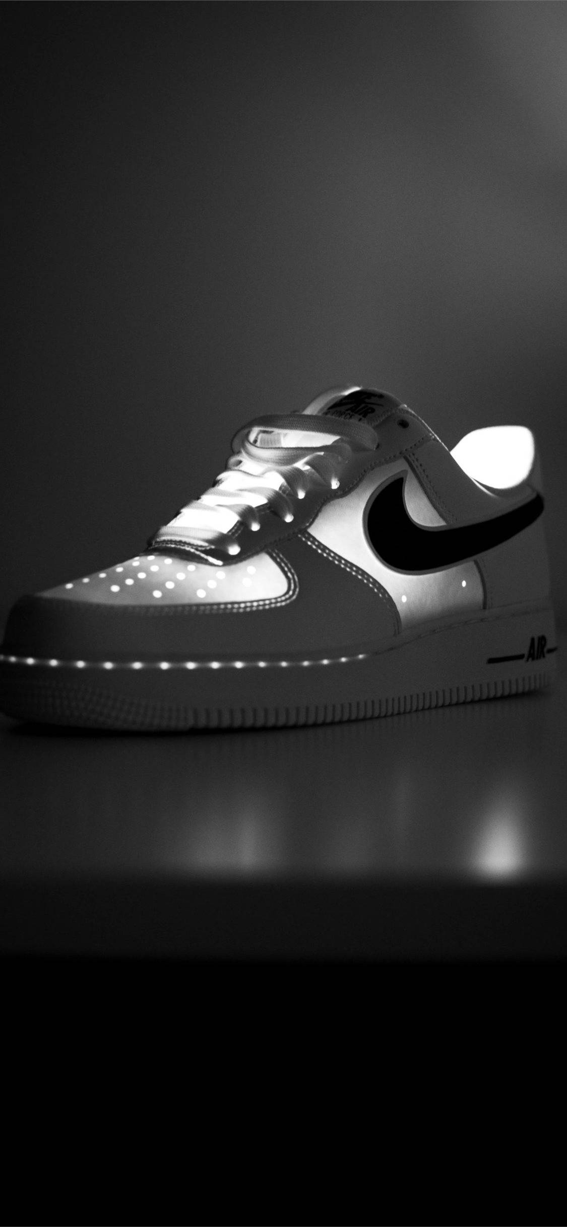 Nike iPhone 桌布 壁紙 — 一雙單色Nike Air Force 1運動鞋，鞋帶和鞋底帶有發光LED燈，放置在反光表面上，背景為深色漸變。