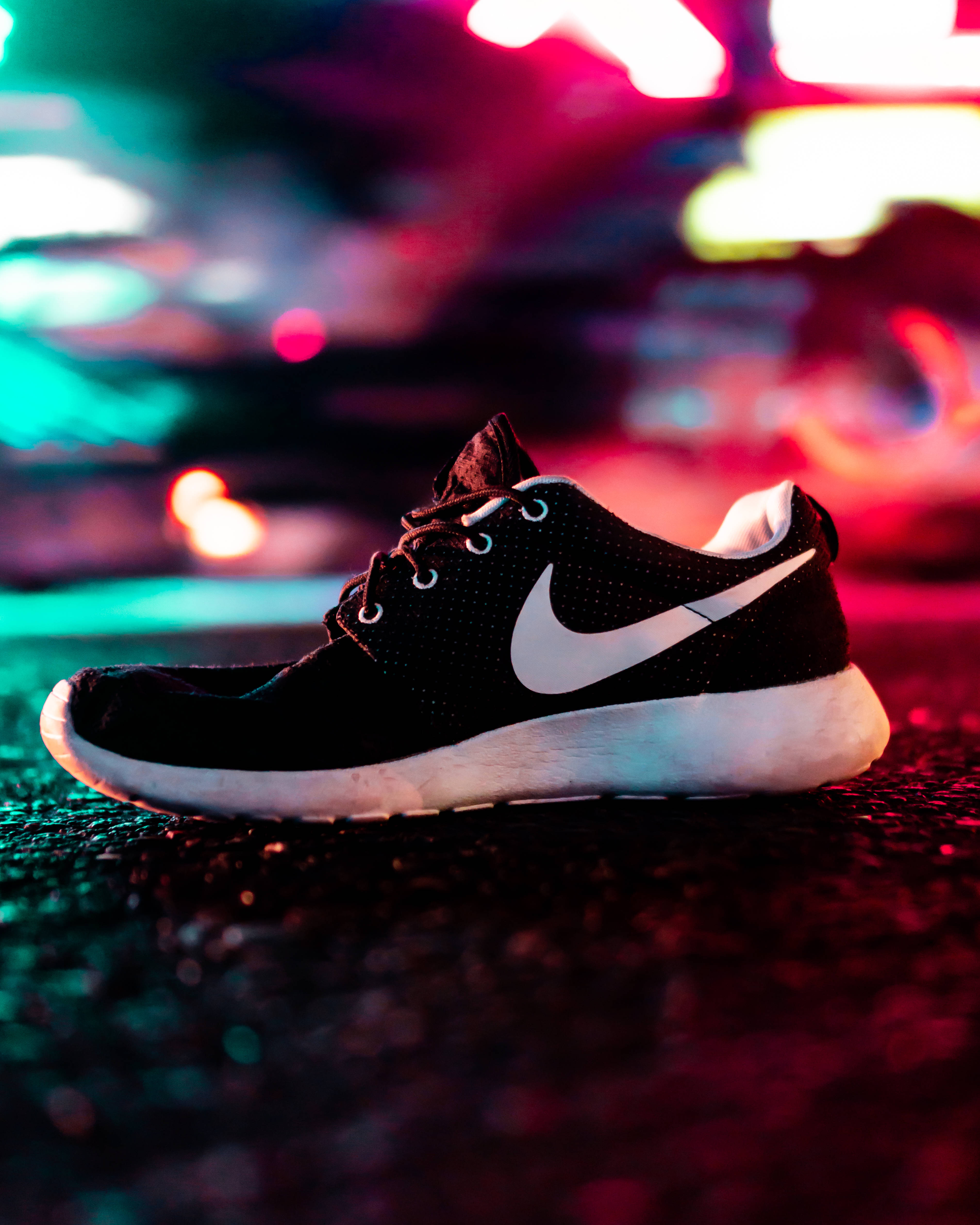 Shoe 桌布 壁紙 — 一雙帶有白色Swoosh的黑色Nike運動鞋在夜間濕漉漉的路面上，在充滿活力的霓虹燈光下休息。