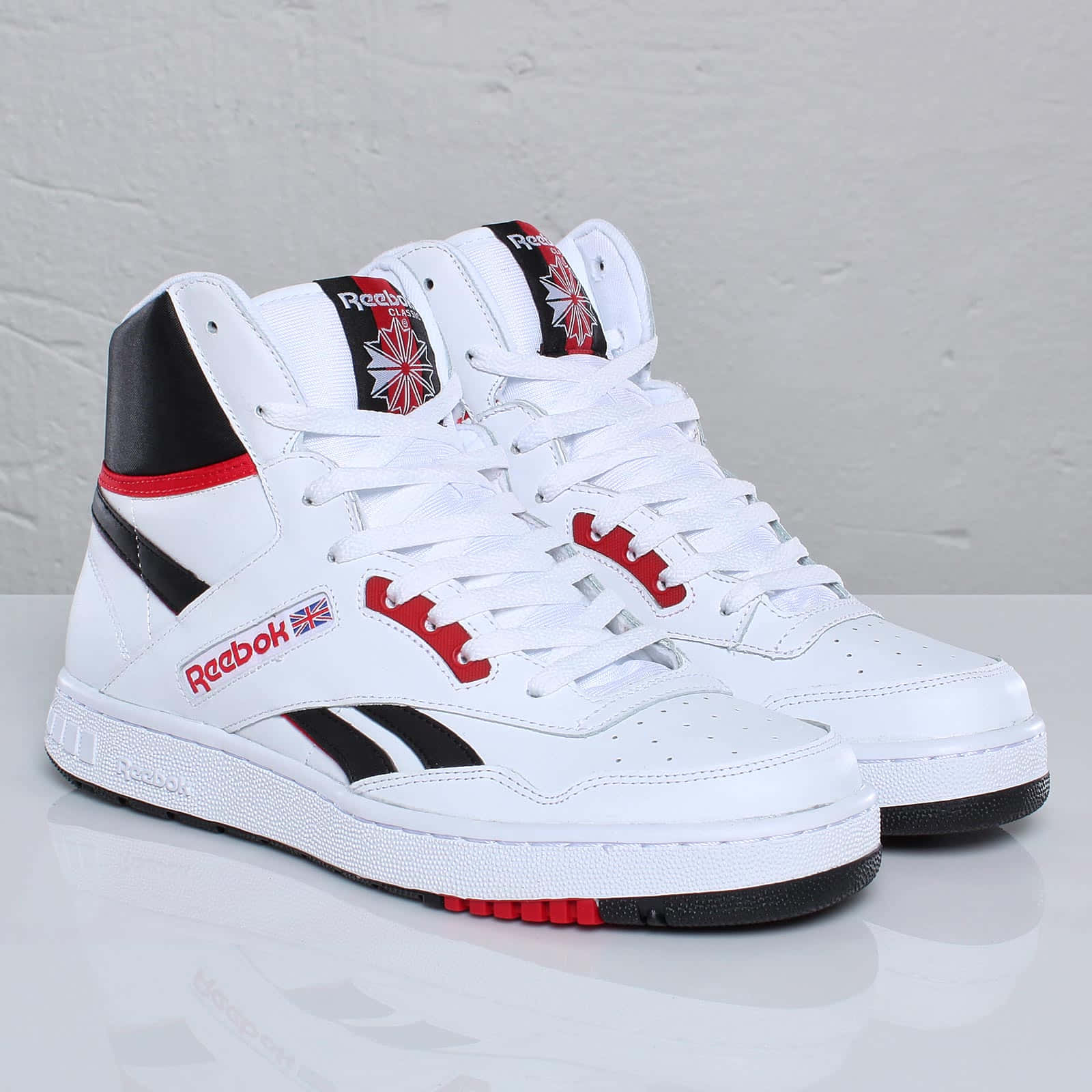 Reebok 桌布 壁紙 — 一雙帶有紅色和黑色點綴的白色高筒Reebok運動鞋，放置在反光表面上，背景是帶紋理的灰色牆壁。