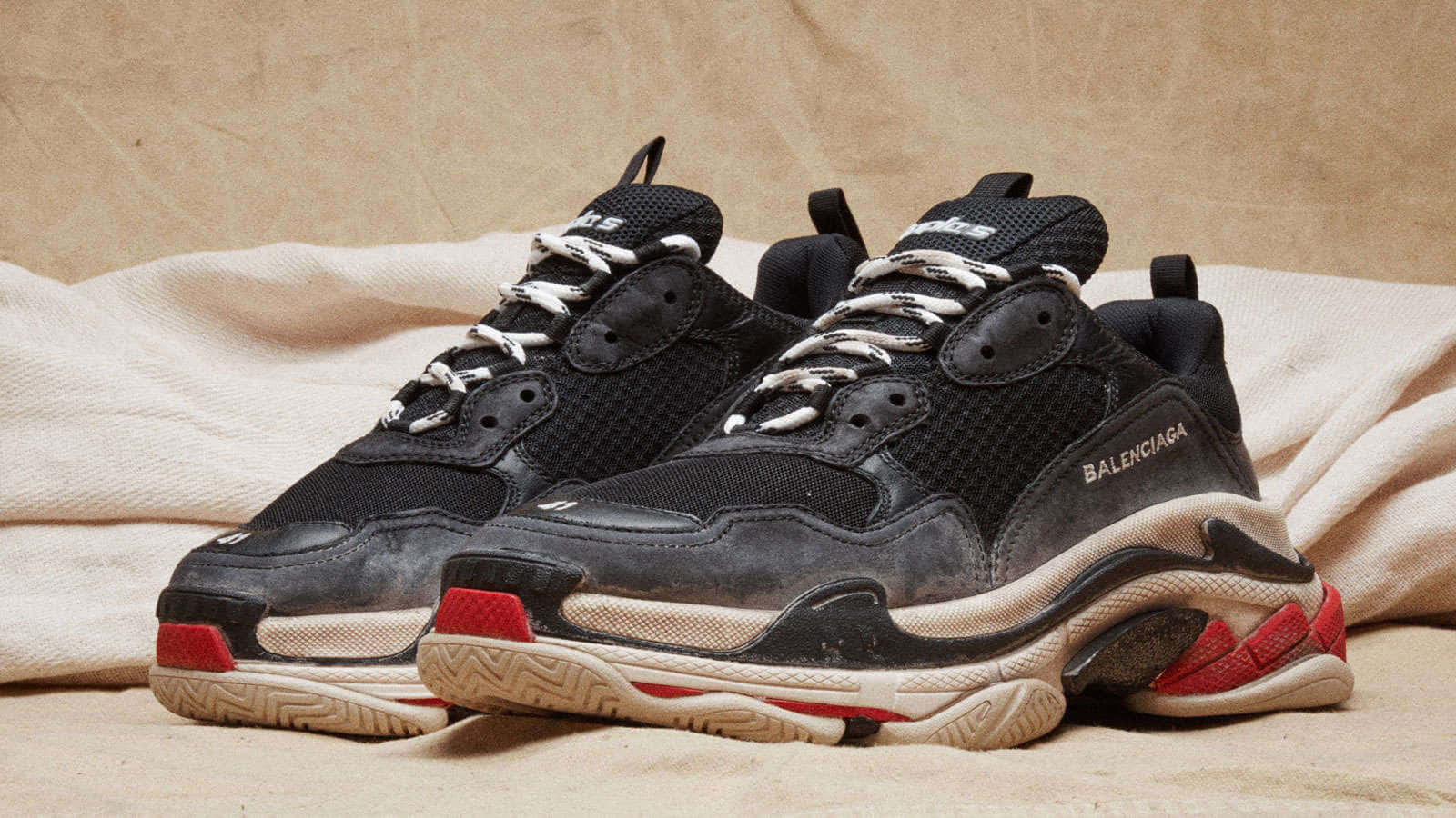 巴黎世家 桌布 壁紙 — 一雙帶有紅色點綴的黑色Balenciaga運動鞋，放在皺巴巴的米色布料上，背景有紋理。
