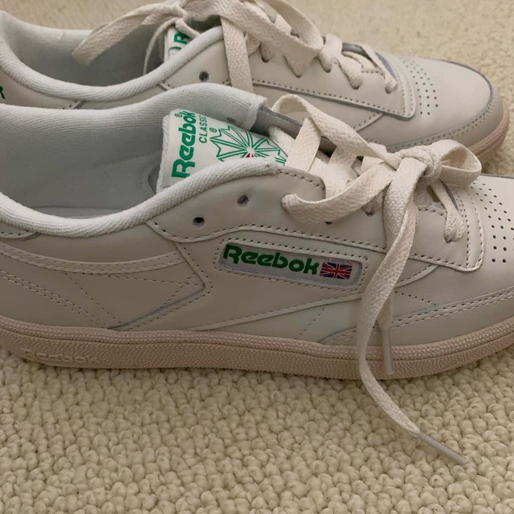 銳步 桌布 壁紙 — 一雙帶有綠色品牌標誌的白色 Reebok Classic 運動鞋特寫，放在有紋理的米色地毯上。