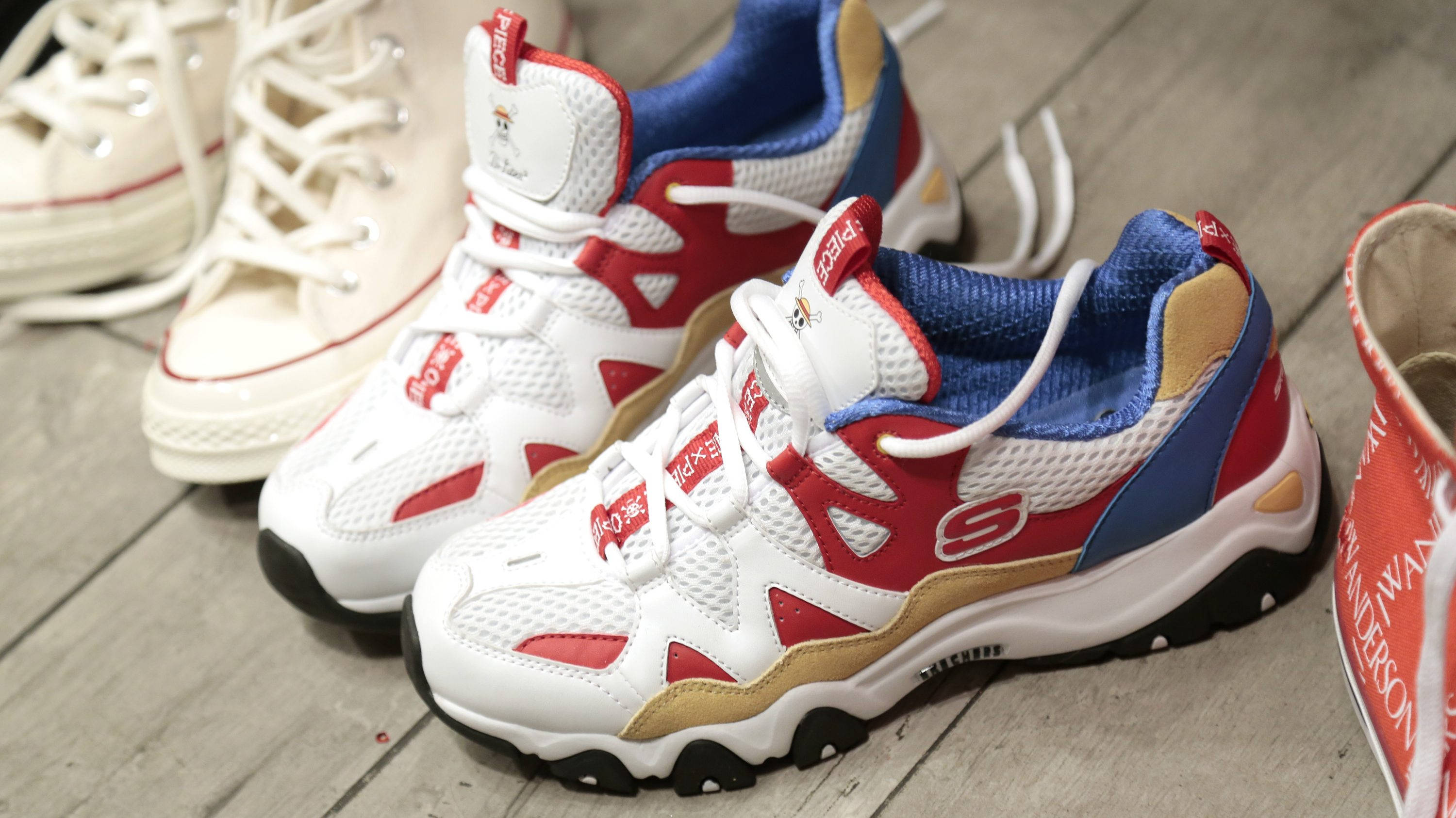 Skechers 桌布 壁紙 — 一雙帶有金色點綴的白色、紅色和藍色厚底運動鞋放在磨損的木地板上，旁邊有一隻米色運動鞋和一個紅色品牌手提包。