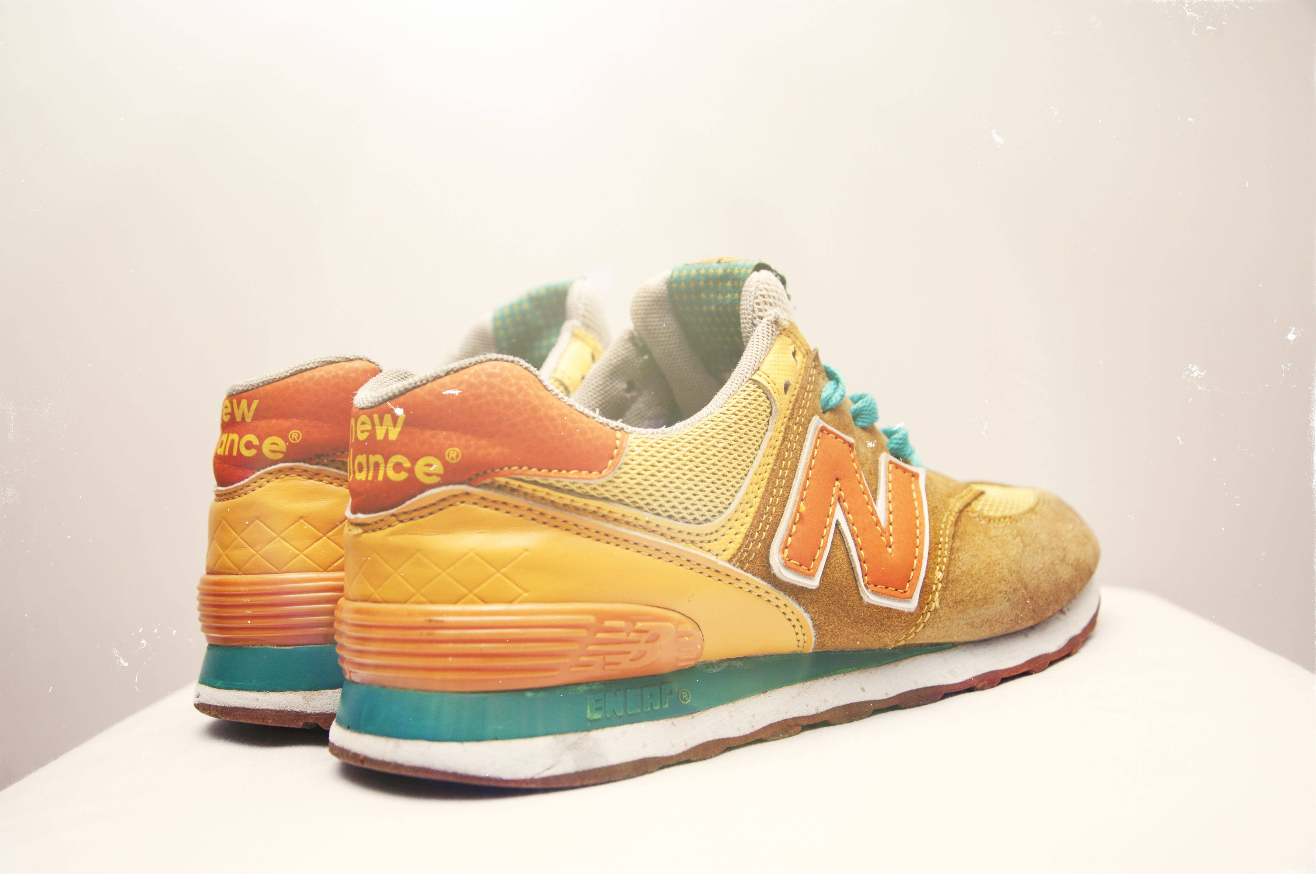 New 桌布 壁紙 — 一雙復古New Balance運動鞋，採用溫暖的橙黃色和藍綠色調，在柔和的白色背景上展示。