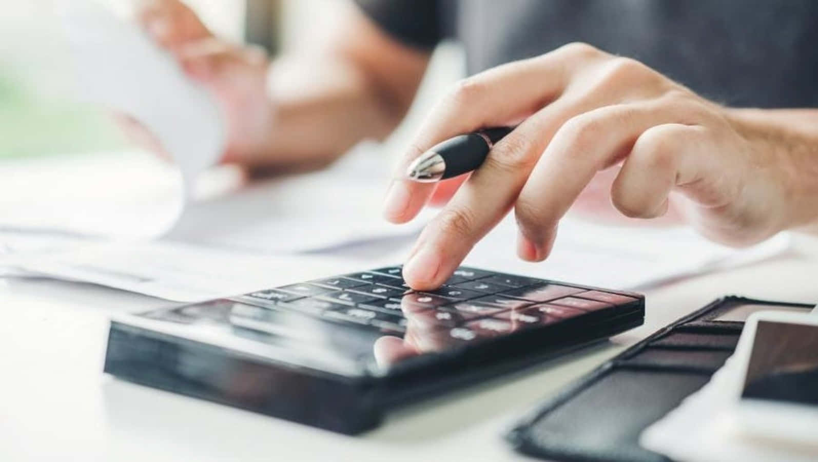 Budgeting 桌布 壁紙 — 一雙手在白色桌上使用黑色計算機，同時握著筆在財務文件上的特寫。