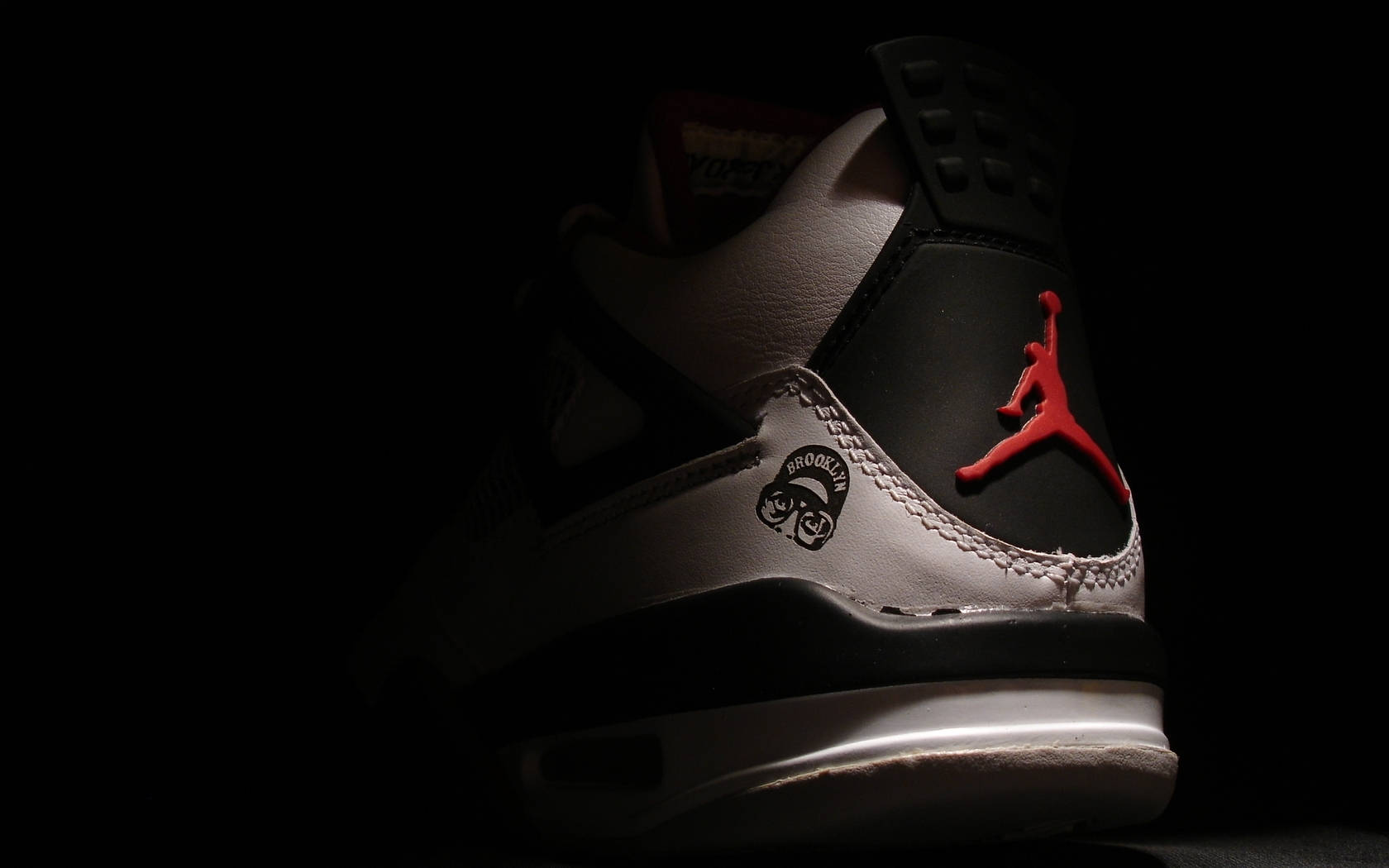 Jordan球鞋 桌布 壁紙 — 一雙時尚的Jordan運動鞋在戲劇性的聚光燈下，在漆黑的背景中閃耀，突顯其紅色標誌和布魯克林補丁。