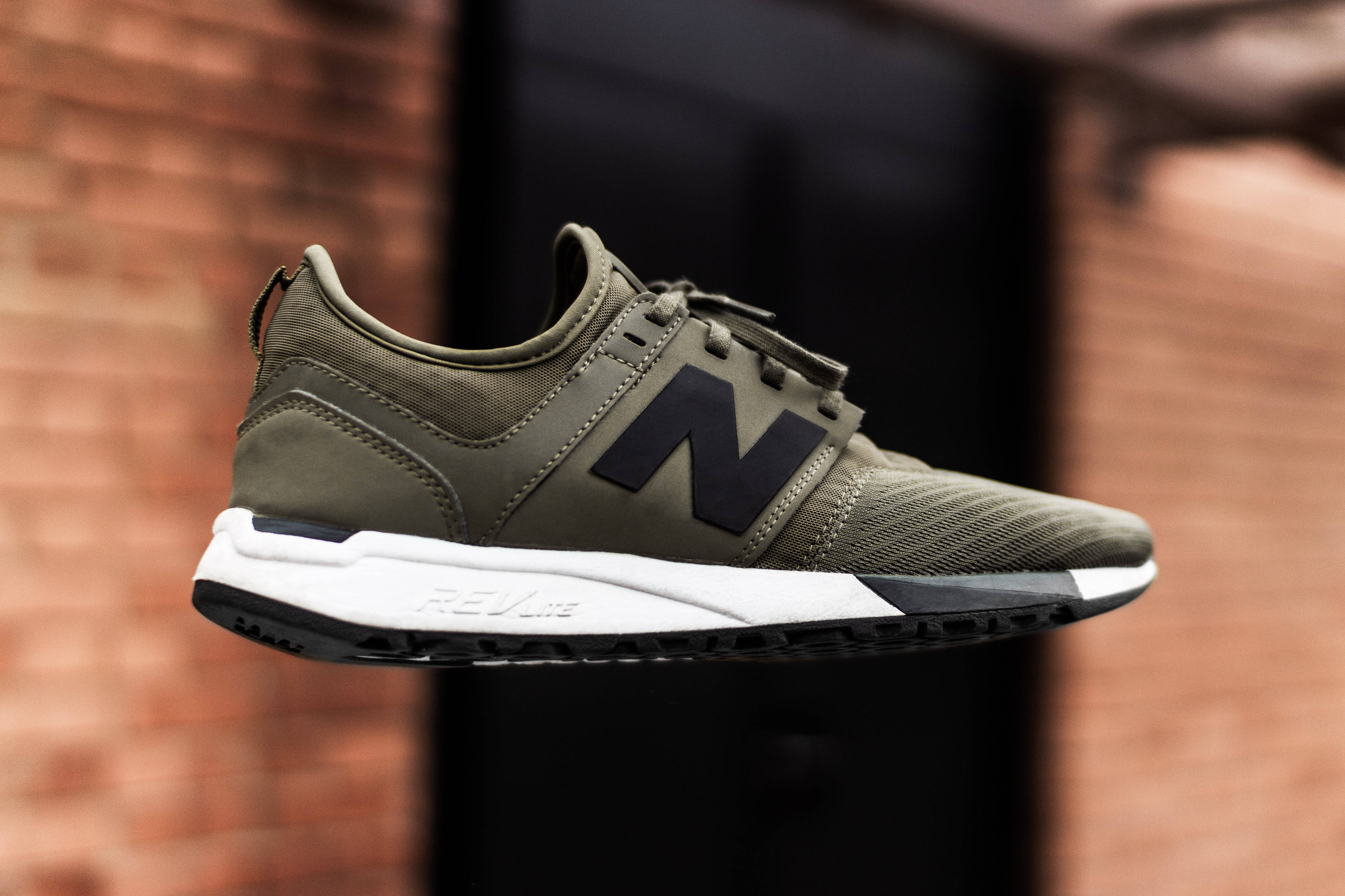New Balance 桌布 壁紙 — 一雙時尚的橄欖綠色New Balance運動鞋，帶有黑色標誌和白色鞋底，漂浮在模糊的磚牆背景上。