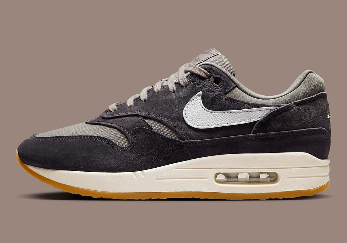 Nike 桌布 壁紙 — 一雙時尚的深灰色 Nike Air Max 1 運動鞋，帶有白色勾勾和橡膠鞋底，側面展示在柔和的棕色背景上。