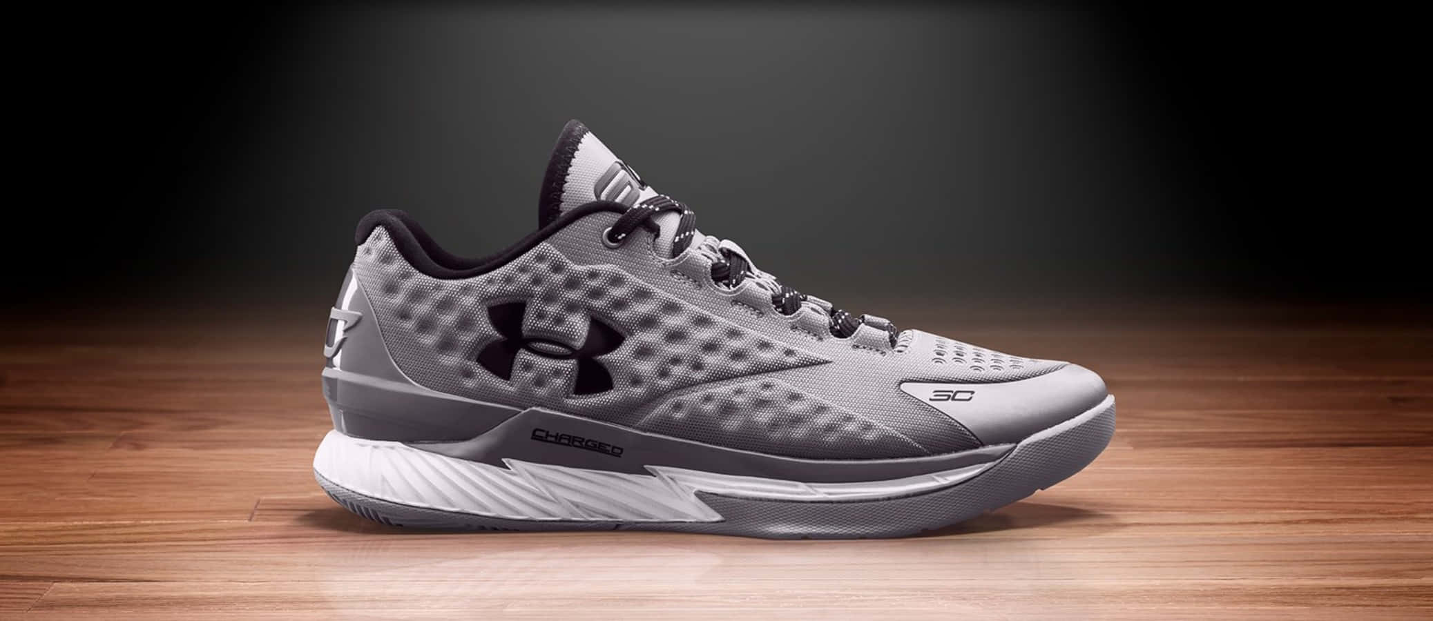 Under Armour 桌布 壁紙 — 一雙時尚的灰色 Under Armour 籃球鞋，帶有黑色點綴，放置在拋光木地板上，背景為深色漸變。