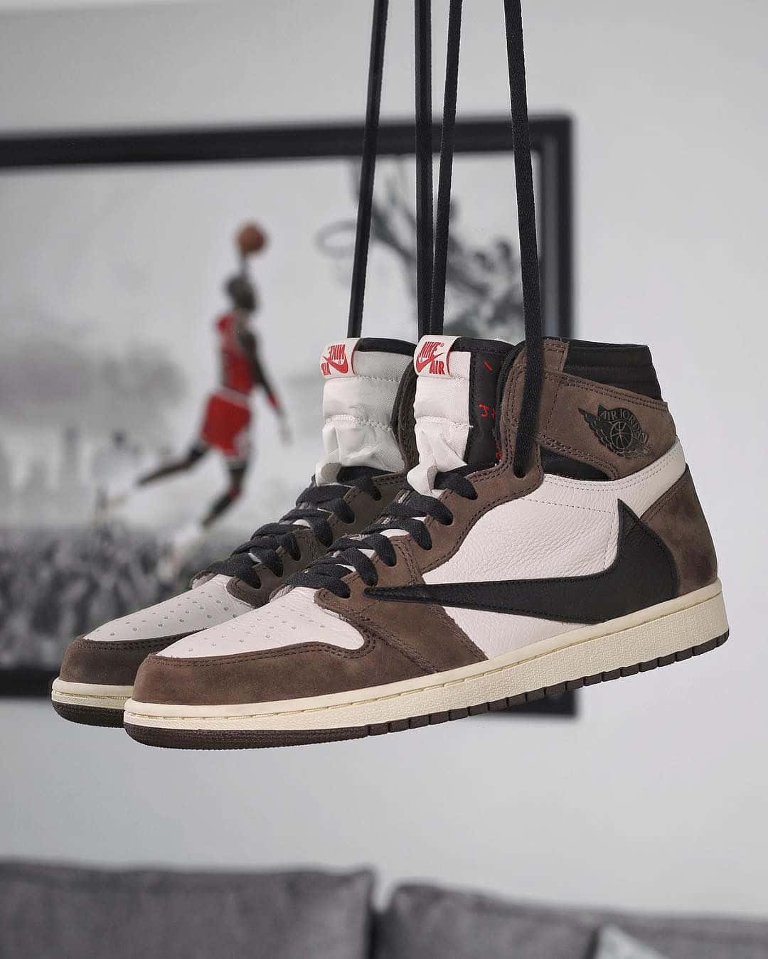 Travis Scott Jordan 1 桌布 壁紙 — 一雙棕白色高筒運動鞋懸掛在鞋帶上，靠著一面牆，牆上掛著一幅裱框的麥可喬登灌籃照片。
