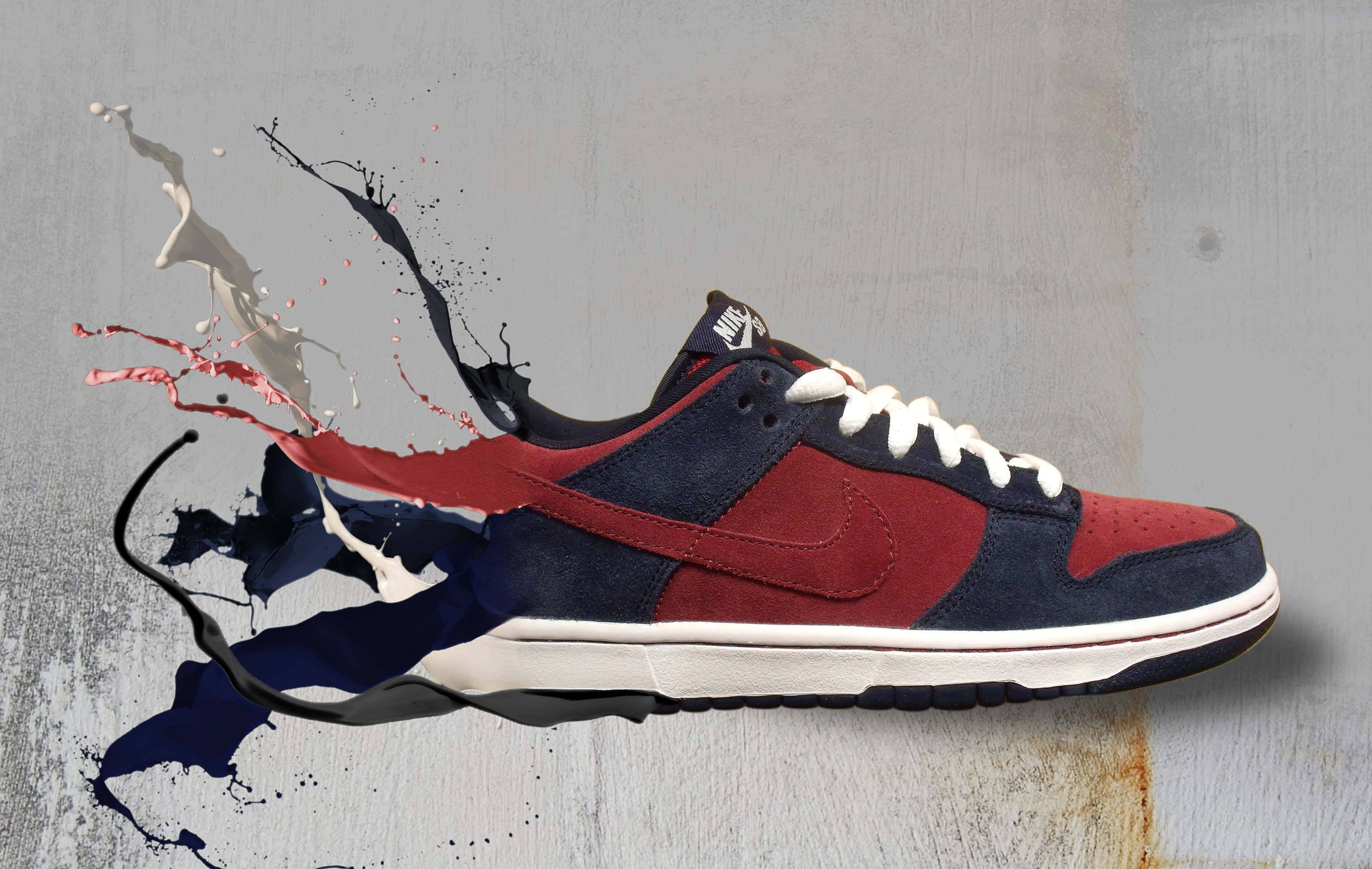 Footwear Design 桌布 壁紙 — 一雙海軍藍和紅色的Nike Dunk運動鞋，在風化的灰色牆壁上爆發出動態的油漆飛濺效果。
