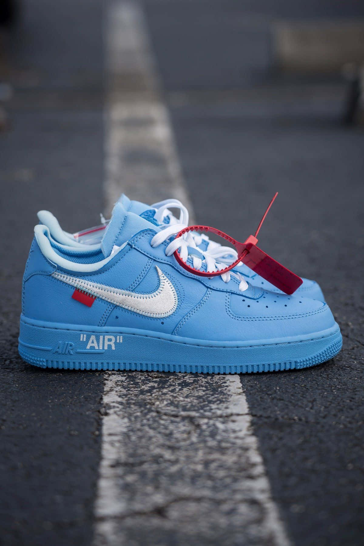 桌布 壁紙 — 一雙淺藍色 Off-White x Nike Air Force 1 運動鞋，帶有紅色標籤，擱在一條有白色標線的裂開柏油路上。
