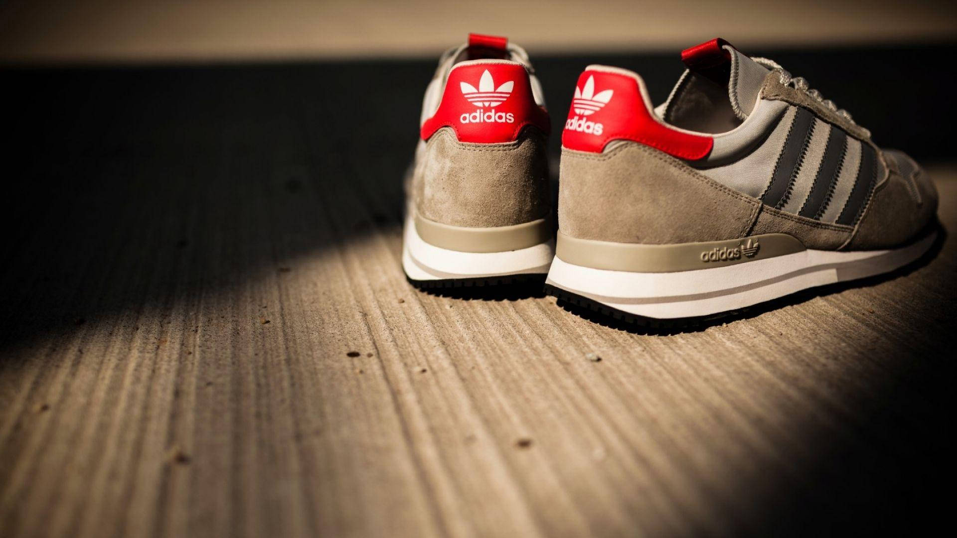 愛迪達 桌布 壁紙 — 一雙灰色 Adidas 運動鞋帶有紅色點綴，在戲劇性聚光燈下放置在木地板上。