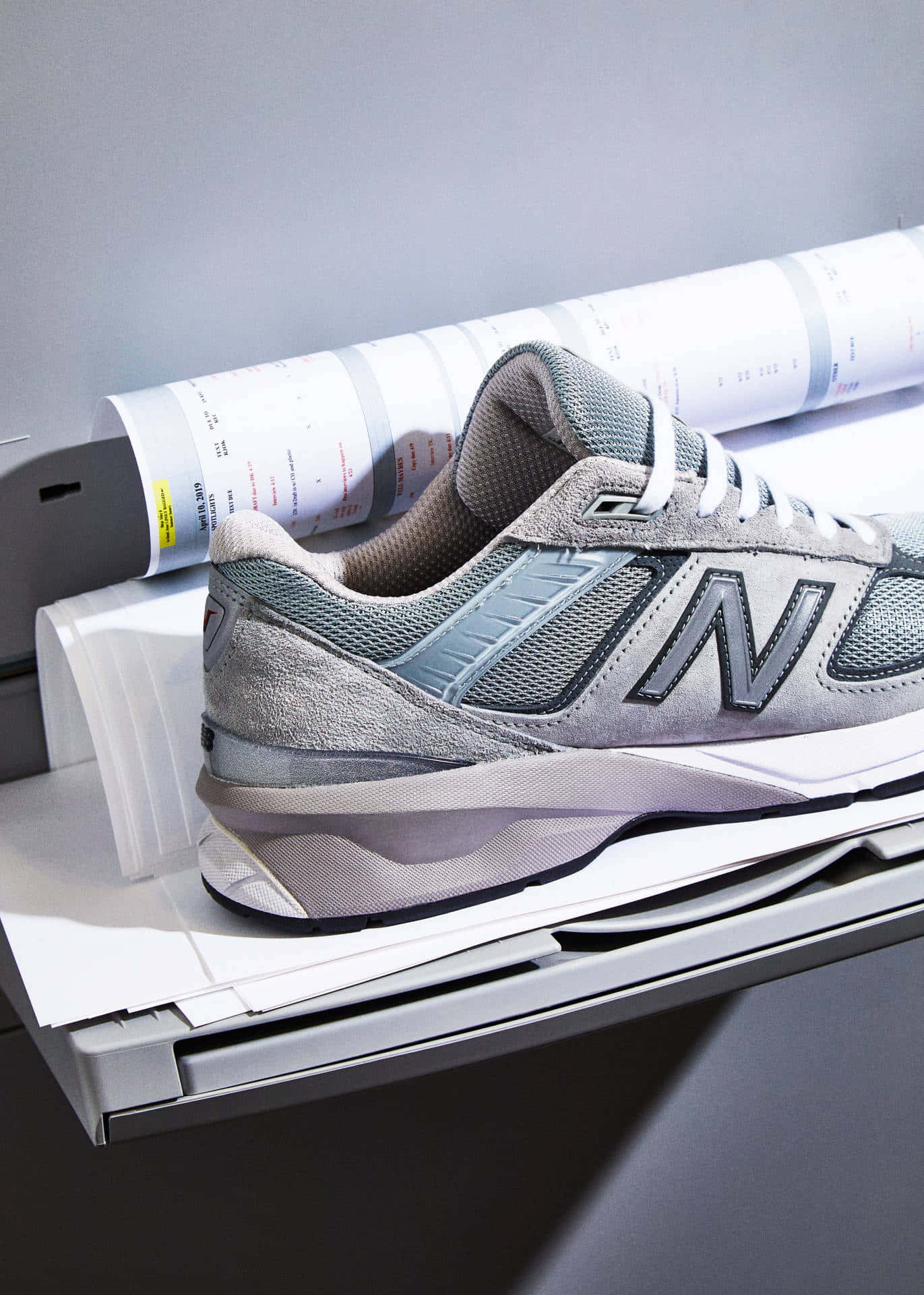 New Balance 桌布 壁紙 — 一雙灰色 New Balance 運動鞋放在印表機托盤上，旁邊有一卷列印好的文件，在柔和的工作室燈光下。